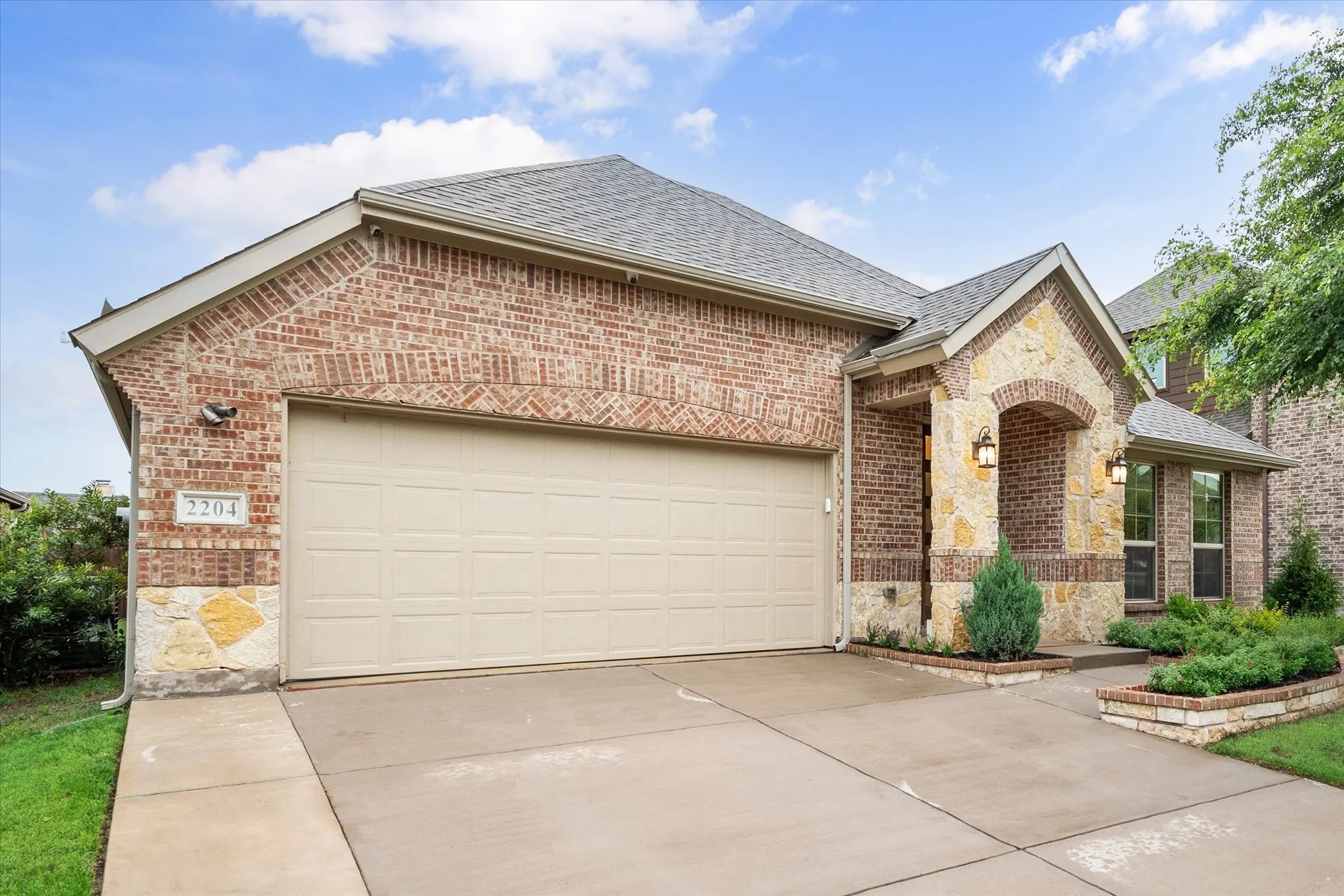 Brick and stone facade, attached two-car garage, covered entry with exterior lighting, and established landscaping