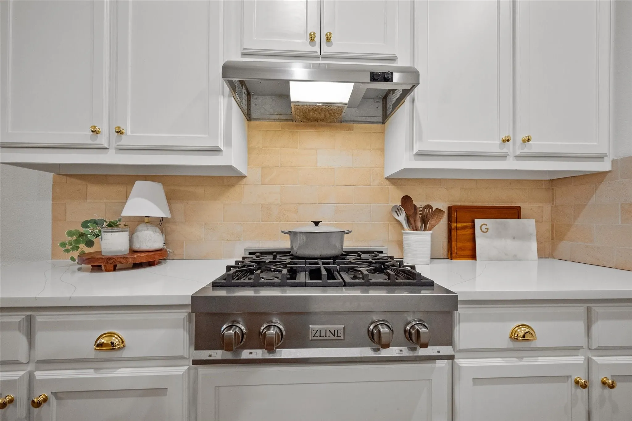 White cabinetry with gold-tone hardware, stainless steel range with griddle, matching range hood, light-toned tiled backsplash, and white countertops