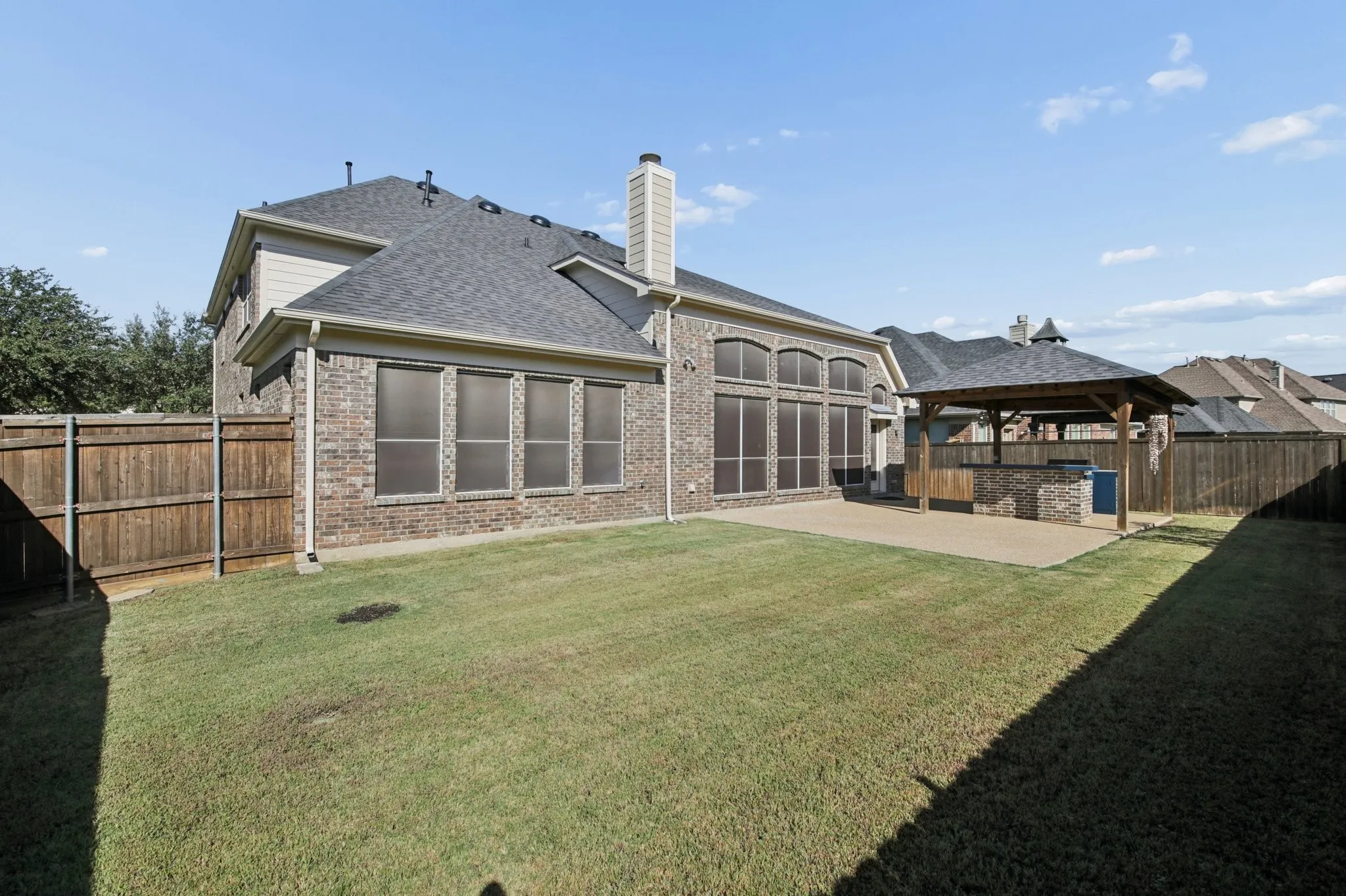 Expansive grassy area featuring a paved patio, a covered outdoor kitchen with a built-in grill, and brick exterior with multiple windows