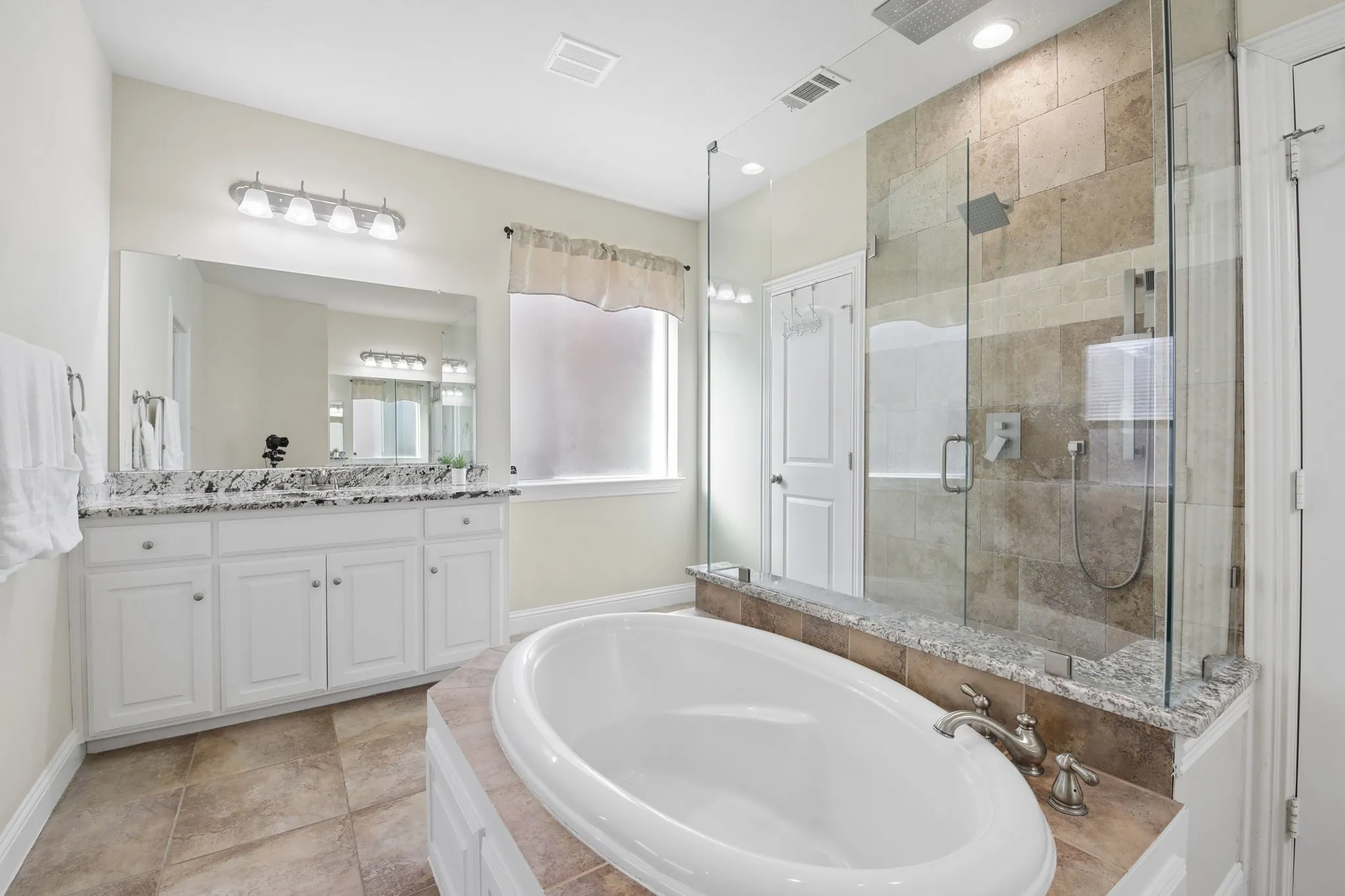 Freestanding soaking tub with deck-mounted faucet, glass-enclosed shower with tile surround, double vanity with granite countertop, and tile flooring