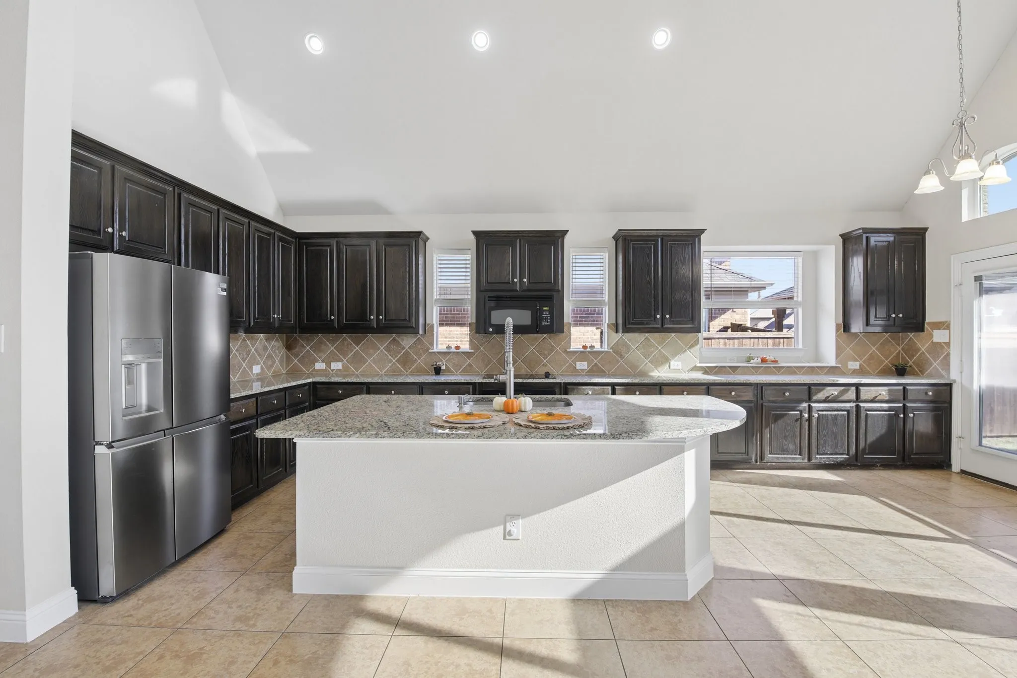 Kitchen featuring an expansive island with a granite countertop, dark wood-finish cabinetry, stainless steel appliances, tiled backsplash, and light-toned tile flooring