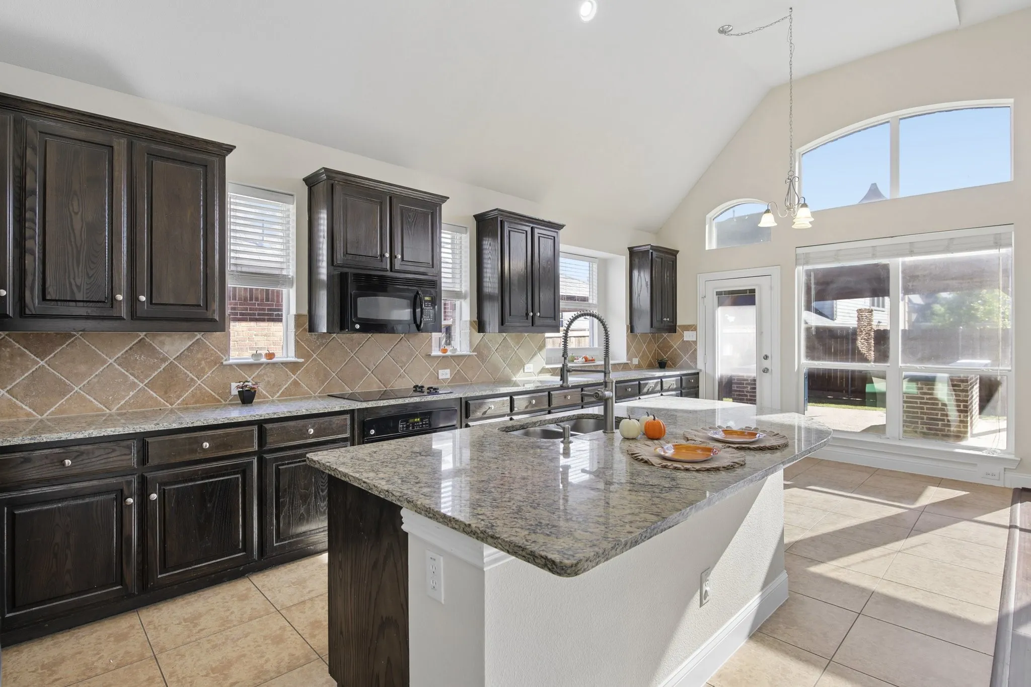Vaulted ceiling kitchen featuring dark wood cabinetry, tile backsplash, stainless steel sink, large center island with stone countertop, and tile flooring