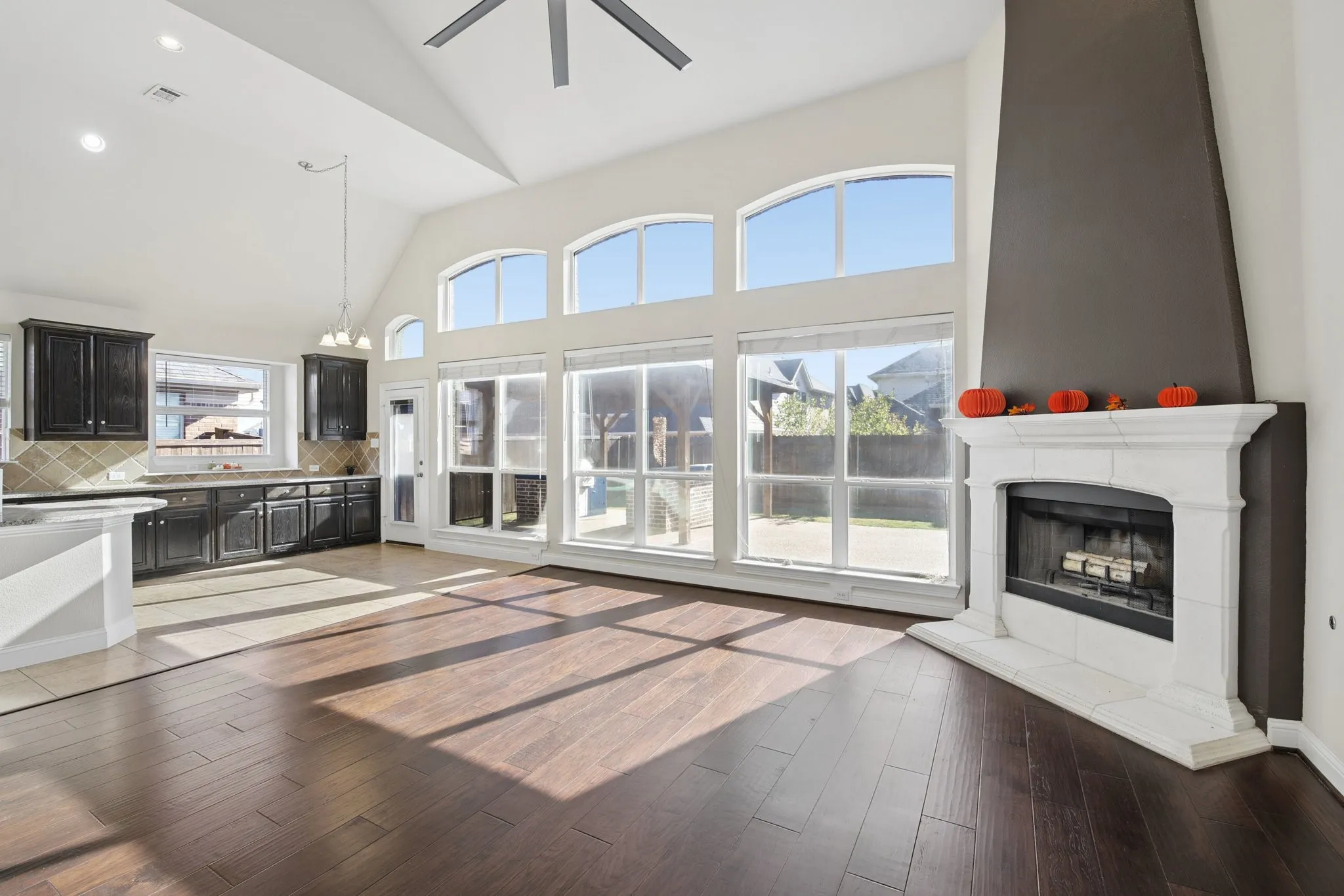 Soaring ceiling with ample natural light, an attached ceiling fan, floor-to-ceiling windows, dark wood-finish flooring, and a stone-clad fireplace