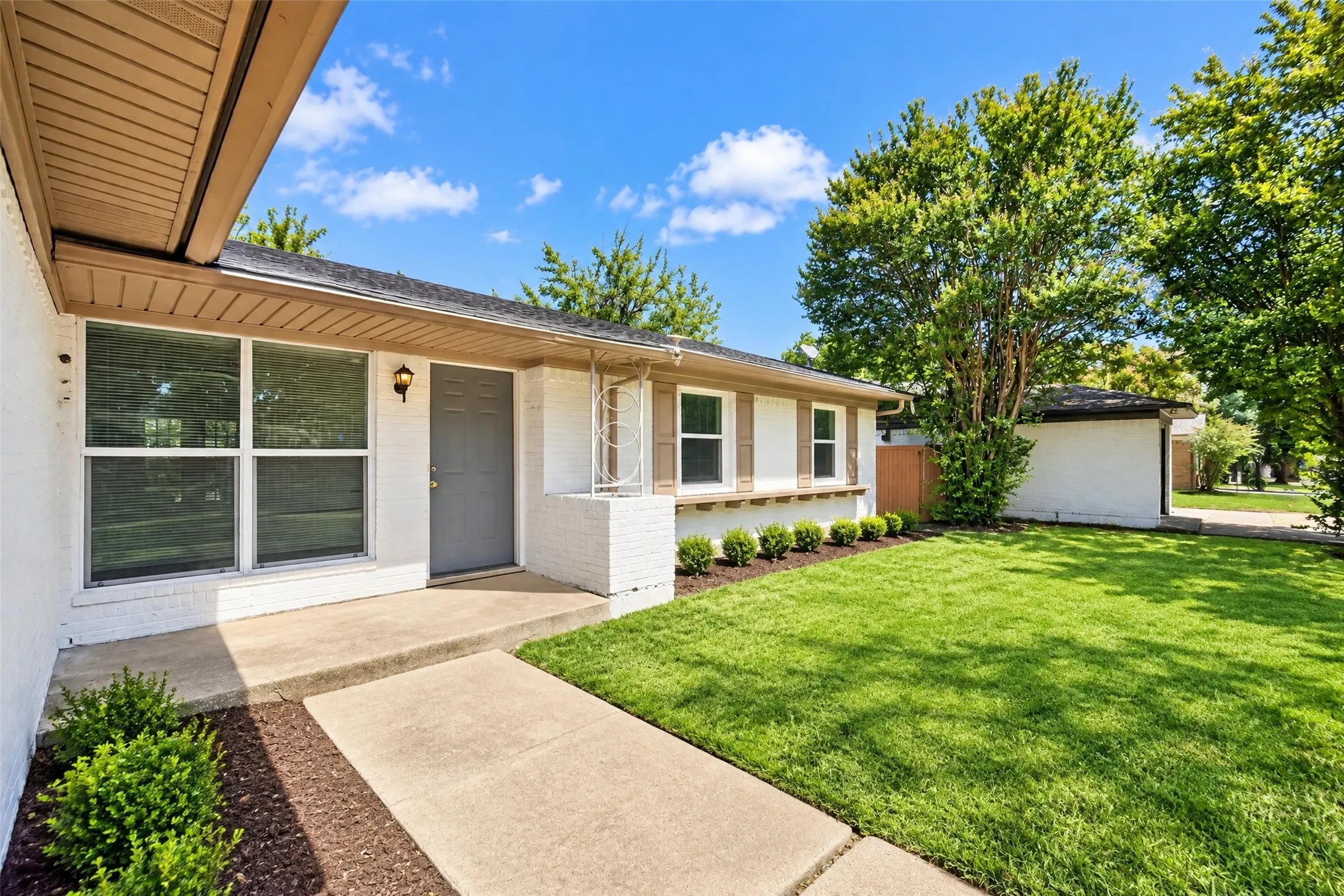 White brick exterior, concrete walkway, vibrant green lawn, landscaping with mature shrubs, and an attached awning