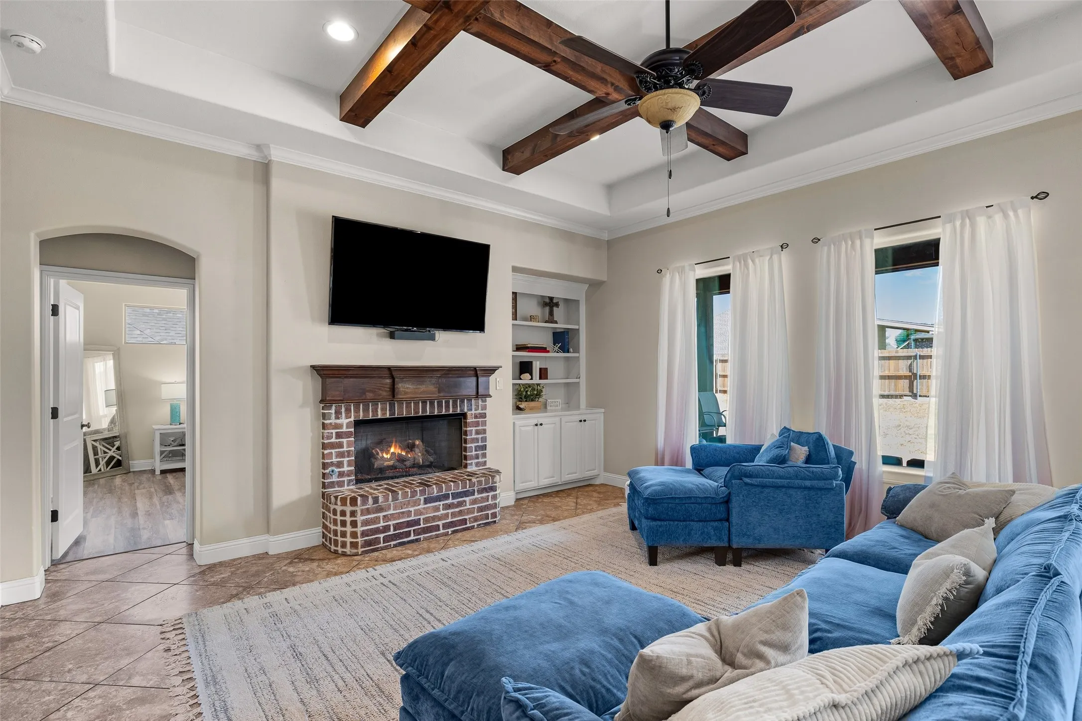 Coffered ceiling with exposed wood beams, brick-surround fireplace with wood mantel, built-in shelving with lower cabinetry, and tile flooring