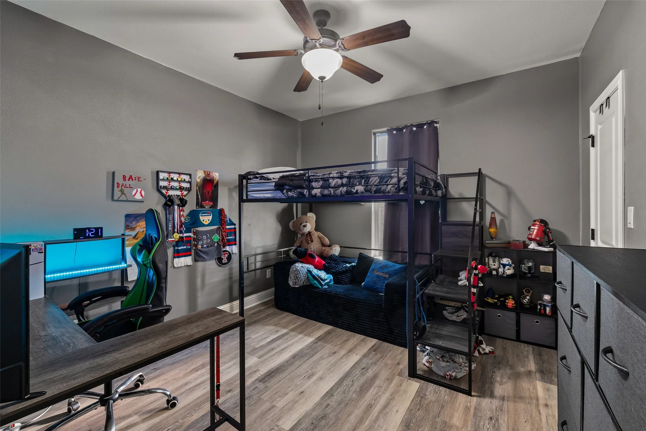 Wood-finish flooring, neutral gray walls, ceiling fan with light fixture, window with drapes, and a white interior door with black hardware