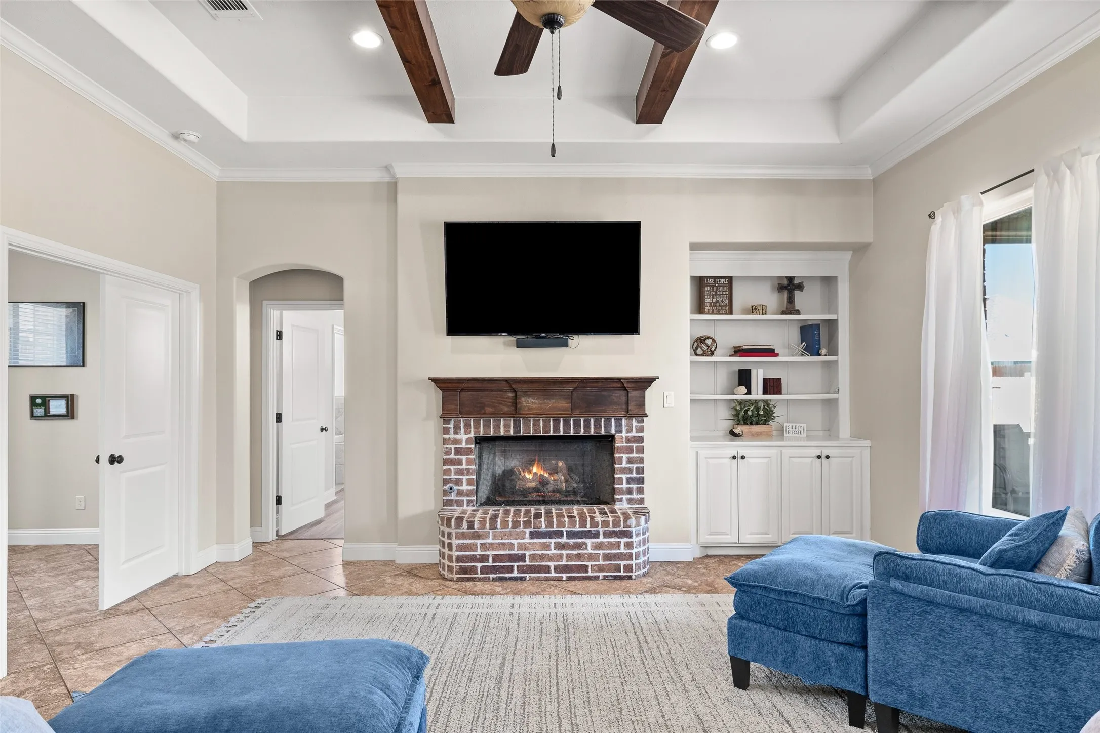 Brick fireplace with a wood mantel, built-in shelving, tray ceiling with exposed wood beams, and a ceiling fan