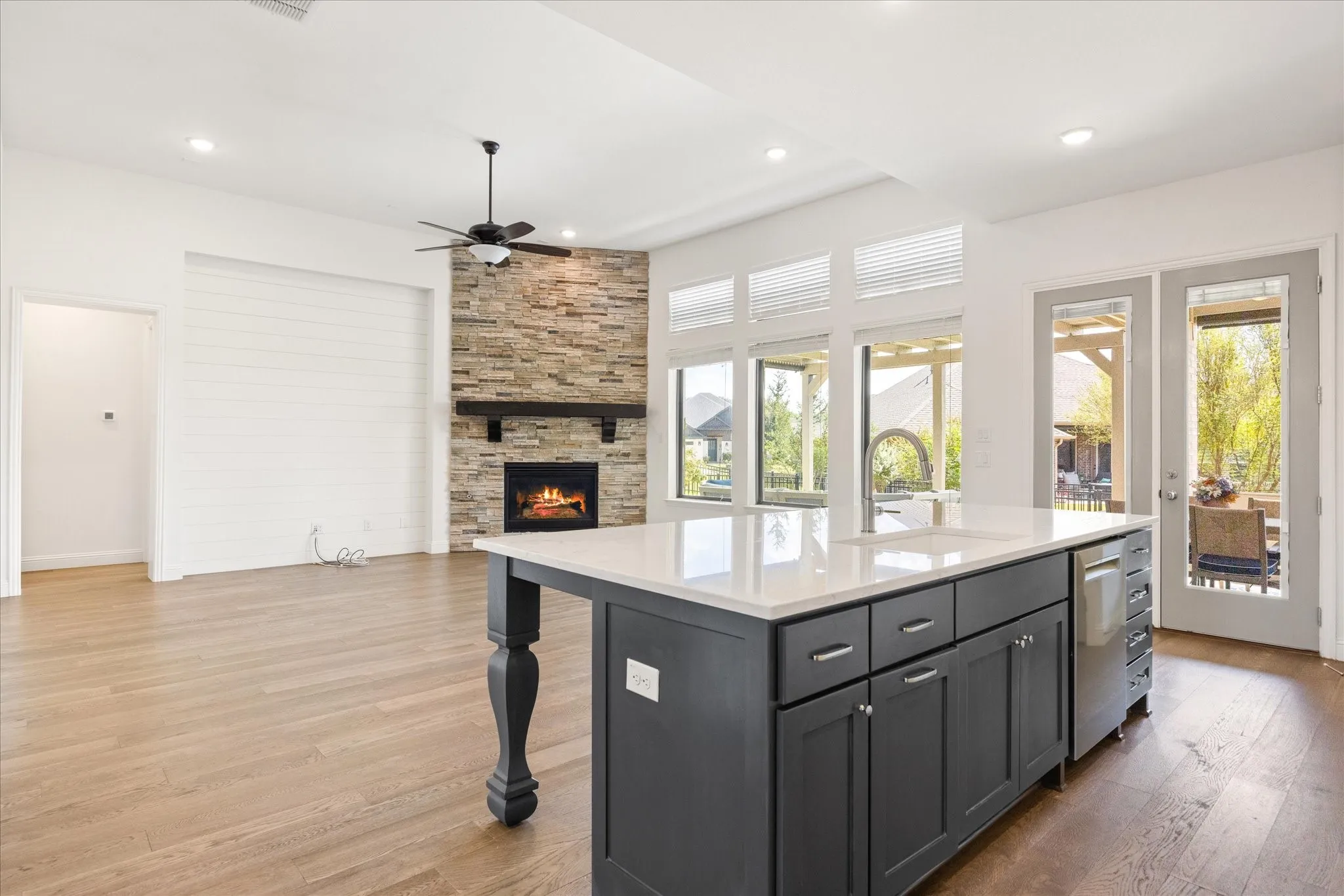 Open-concept living space with wood-finish flooring, a stone-clad fireplace with a dark wood mantel, and large windows
