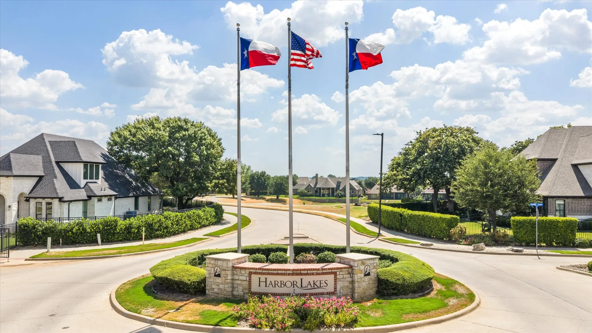 Stone-clad monument with "Harbor Lakes" inscription, three flagpoles, manicured hedges, and paved roadways