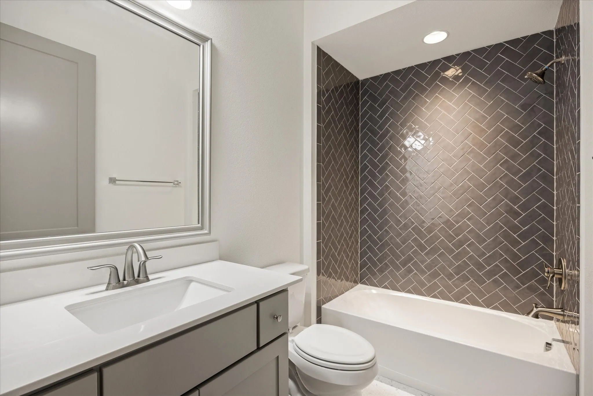 Bathroom featuring a sleek gray vanity, white countertop, undermount sink with brushed nickel faucet, and a shower-tub combination with dark gray herringbone tile surround