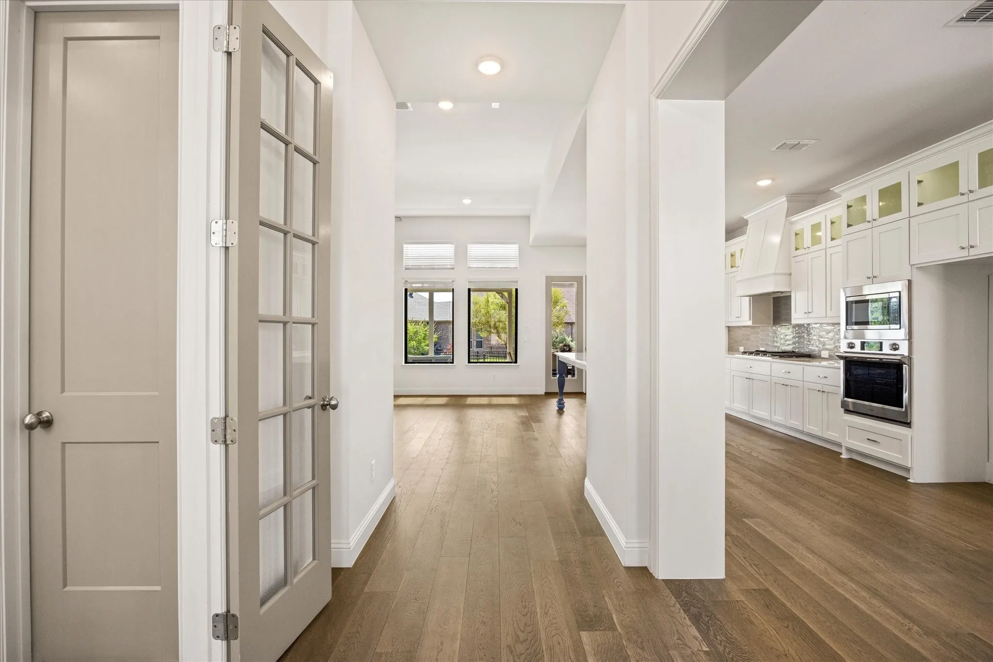 Wood-finish flooring throughout, white baseboards, recessed lighting, white kitchen cabinetry with glass paneling, and stainless steel built-in appliances