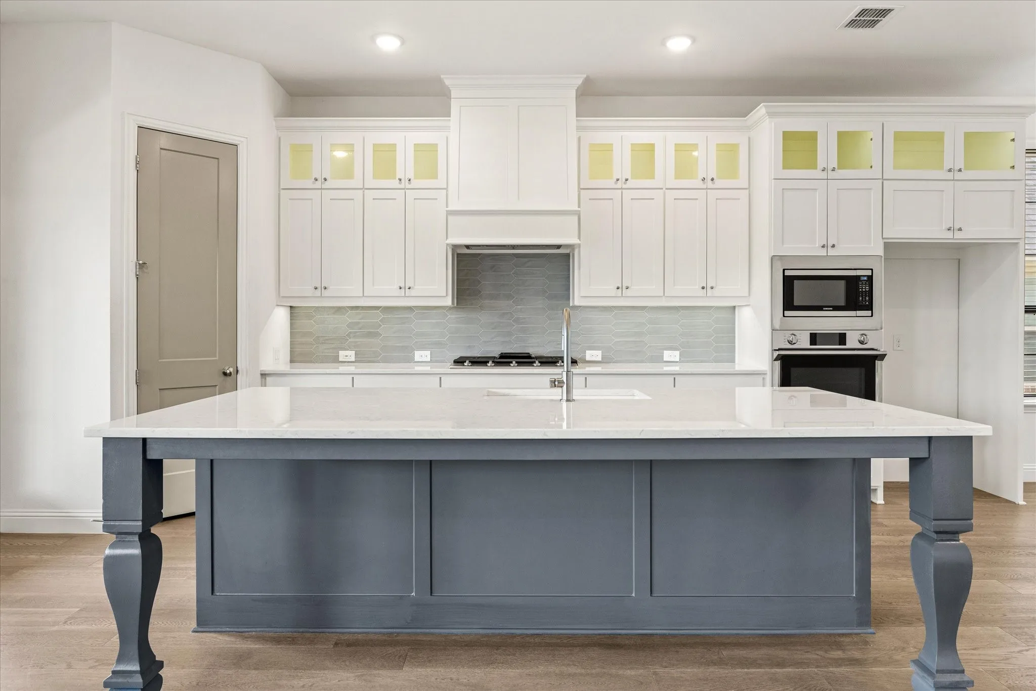 Kitchen featuring a large central island with a light-toned countertop, white cabinetry with glass-front uppers, a patterned tile backsplash, and stainless steel appliances