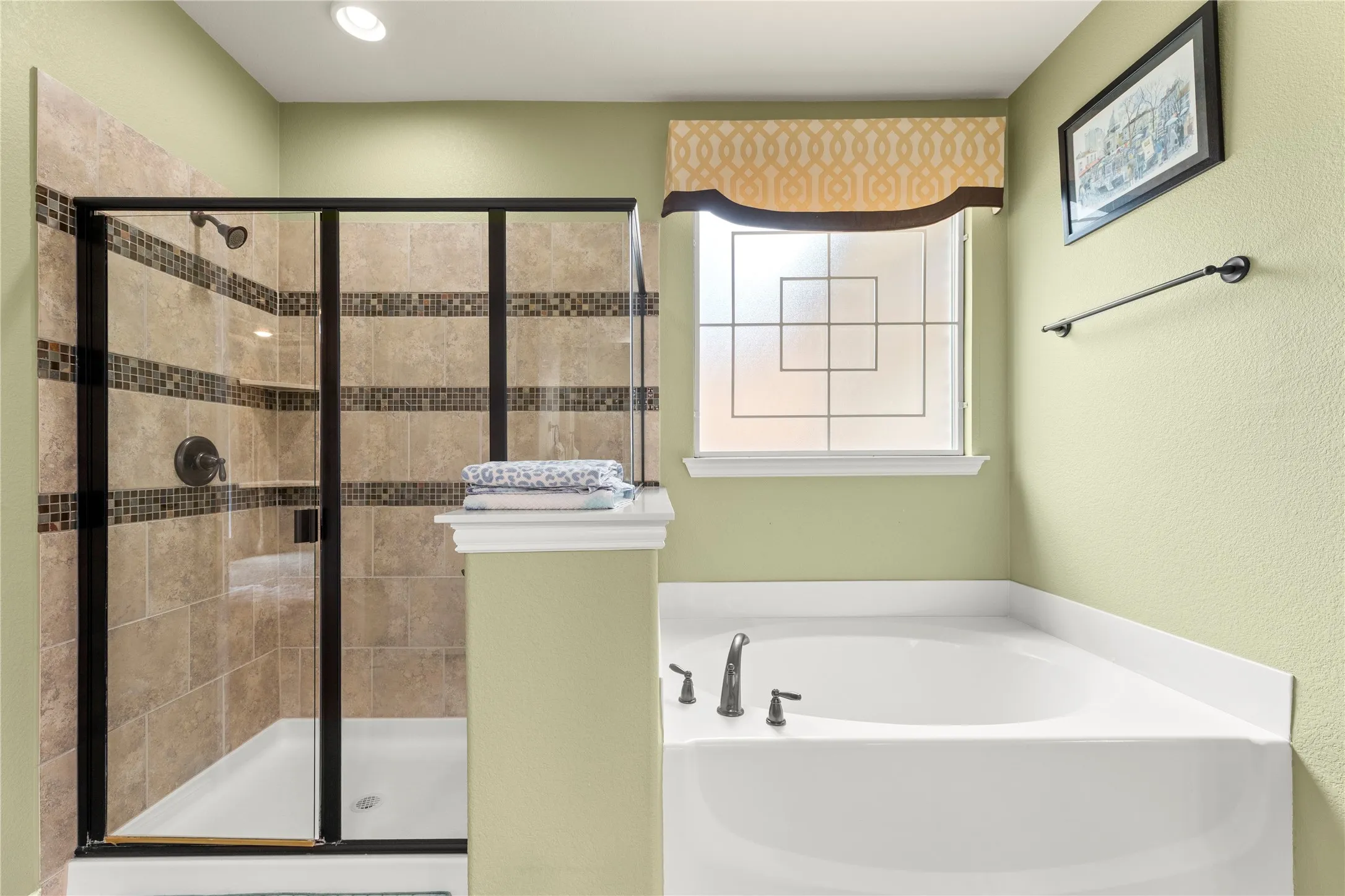 Spacious bathroom featuring a glass-enclosed shower with tile surround and decorative mosaic accents, a built-in soaking tub with dark-finish fixtures, and a privacy window with patterned glass
