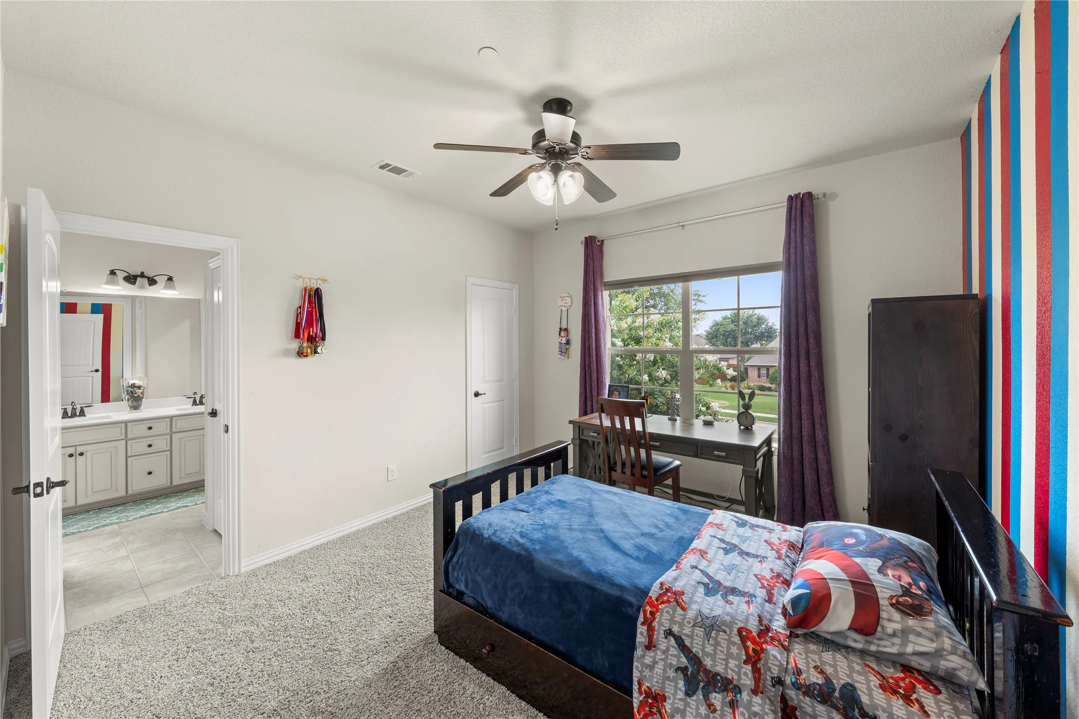 Carpeted bedroom featuring a large window, ceiling fan, and direct bathroom access with a double vanity and tile flooring