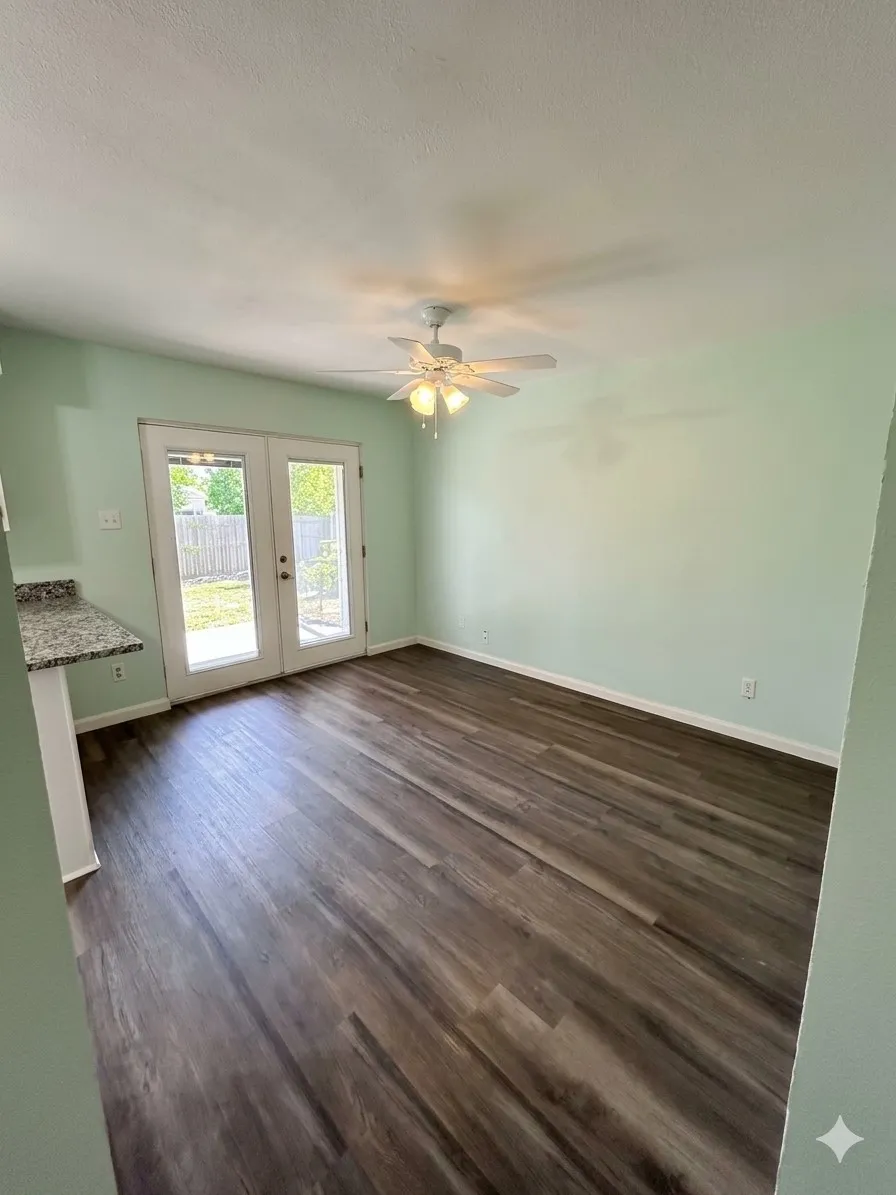 Wood-finish flooring, white ceiling fan with light fixture, light green wall paint, white French doors, and a granite-finish countertop
