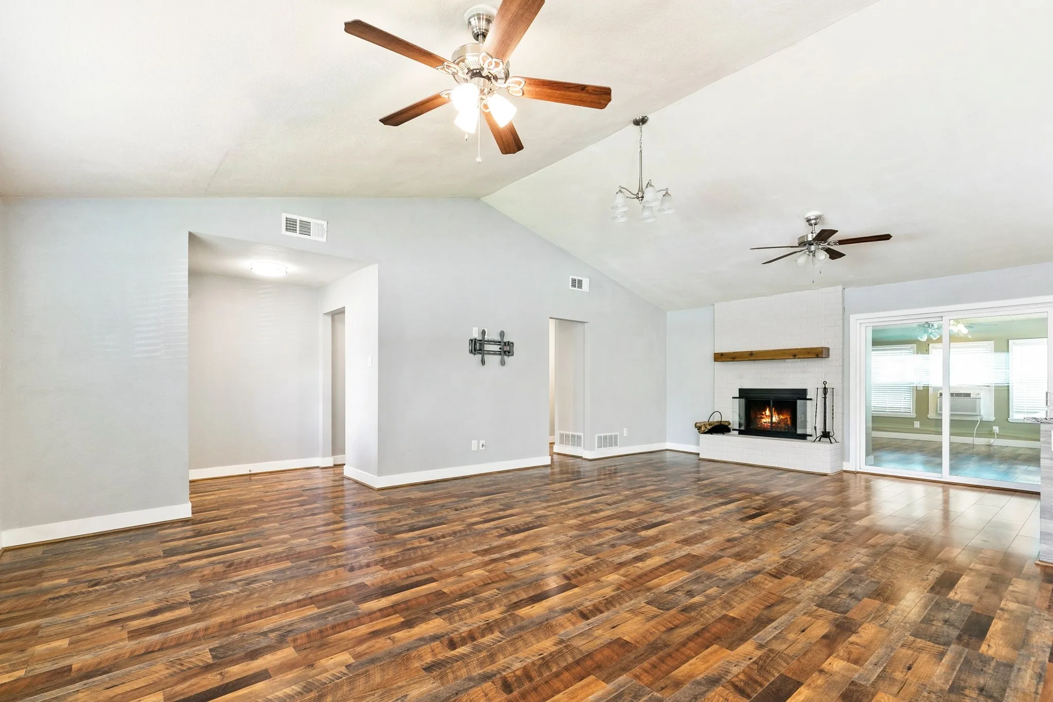 Vaulted ceilings, wood-finish flooring, white brick fireplace with wood mantel, multiple ceiling fans, and sliding glass doors
