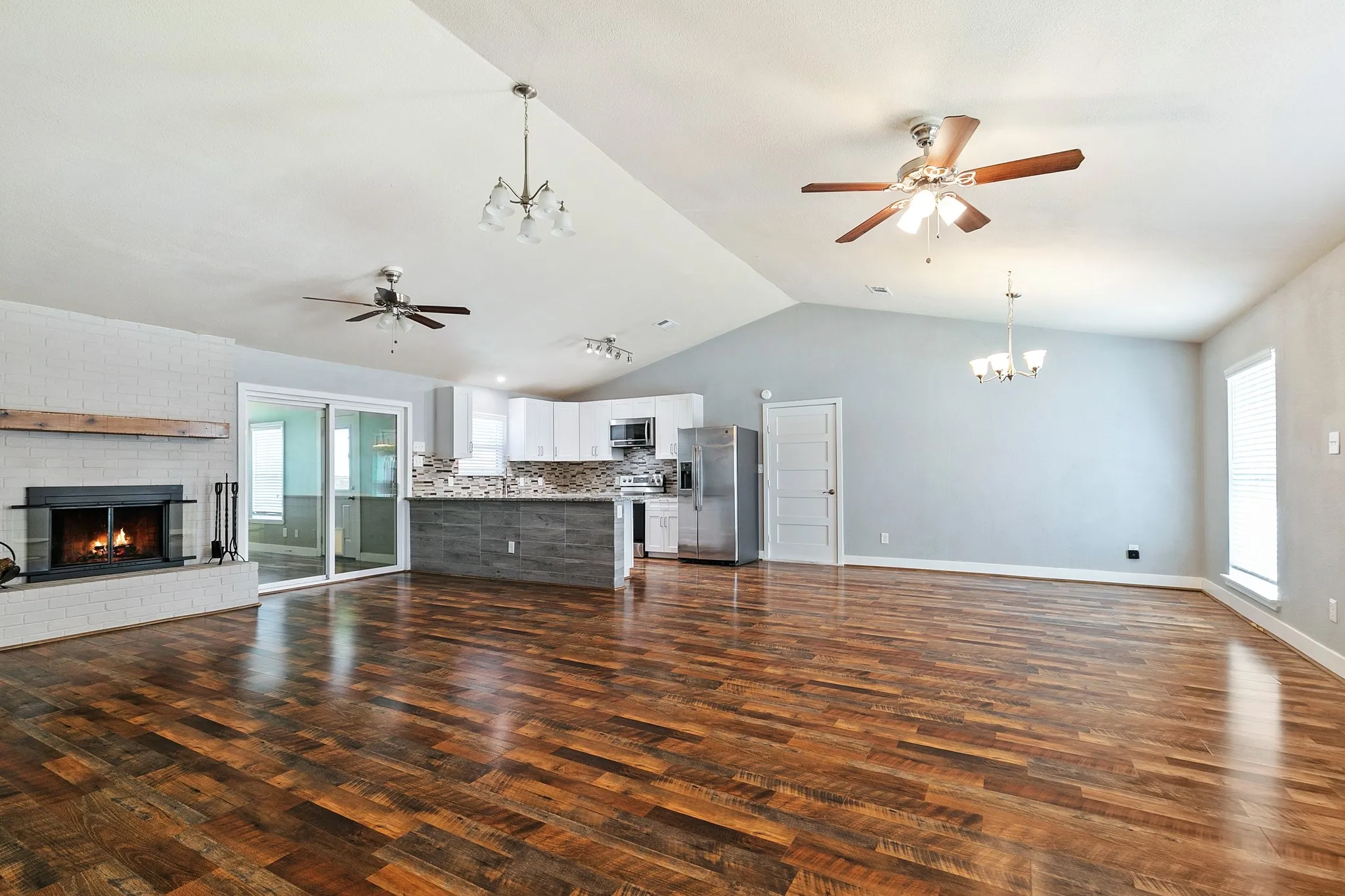 Vaulted ceiling with multiple ceiling fans and chandeliers, wood-finish flooring, white brick fireplace with a wood mantle, and a kitchen featuring white cabinetry and stainless steel appliances