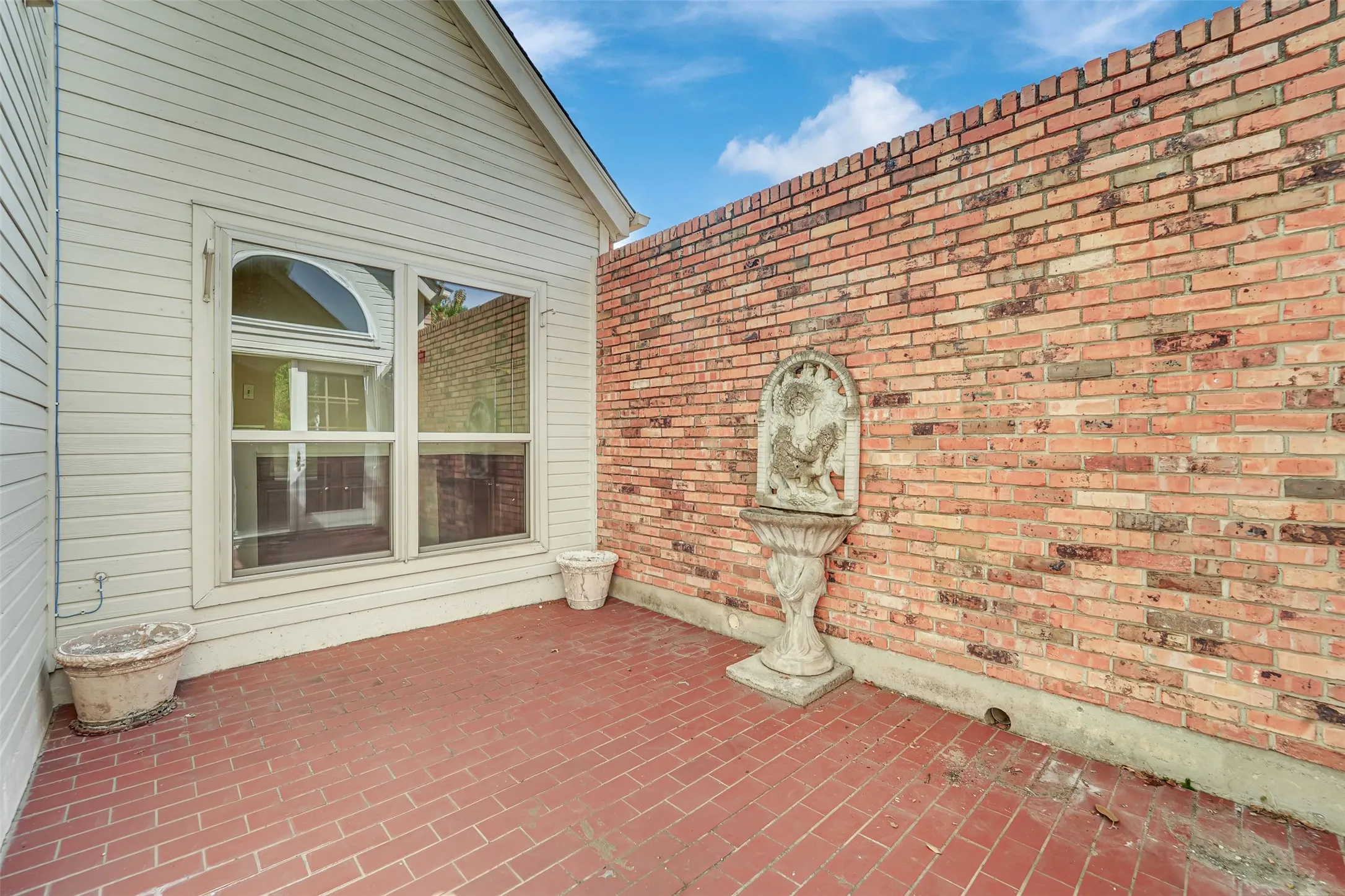 Paved patio area featuring brick flooring, a tall brick privacy wall, and a large multi-paned window with an arched transom