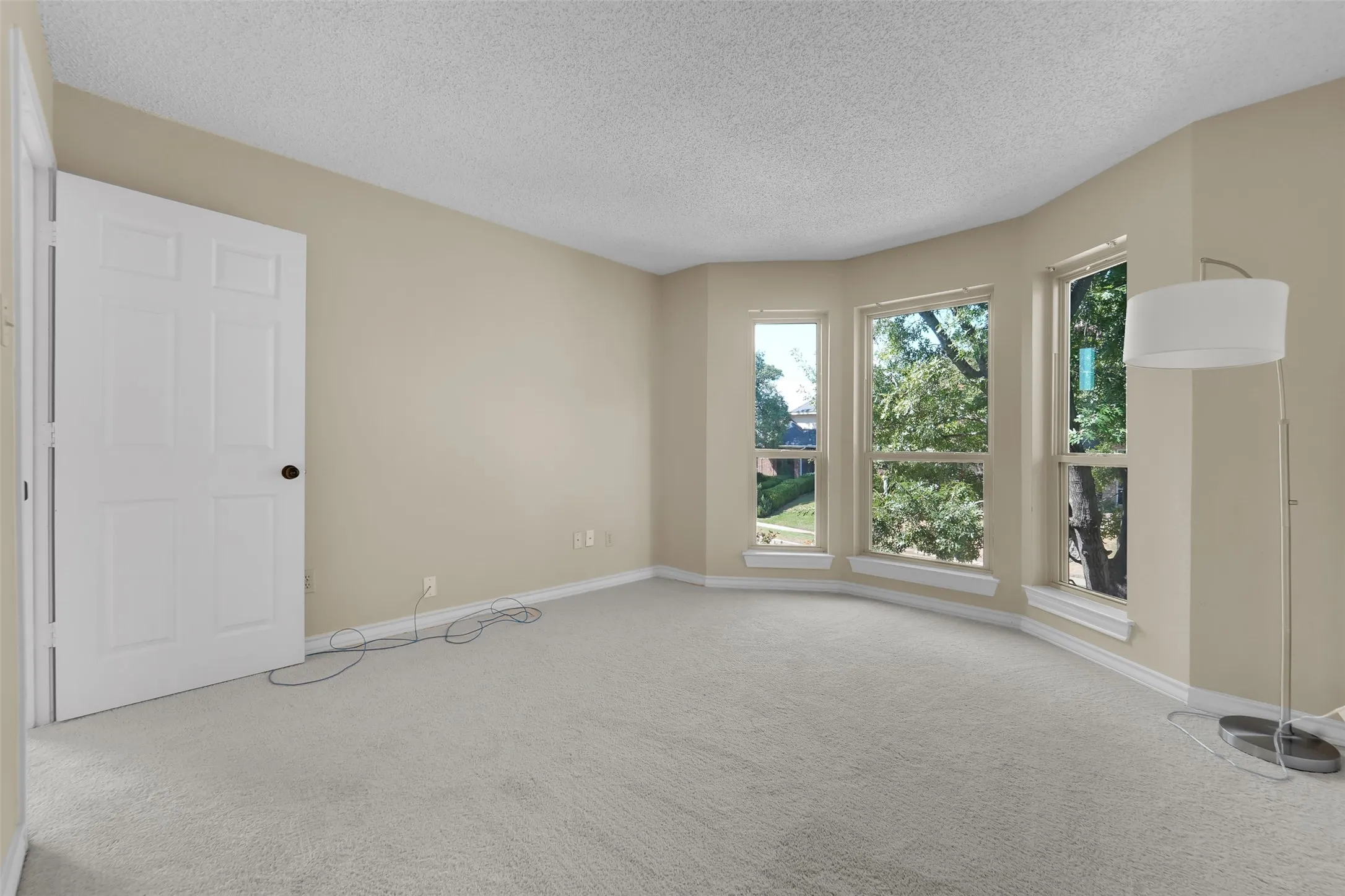 Bay window with three large windows, textured ceiling, light-colored carpeting, and neutral wall paint