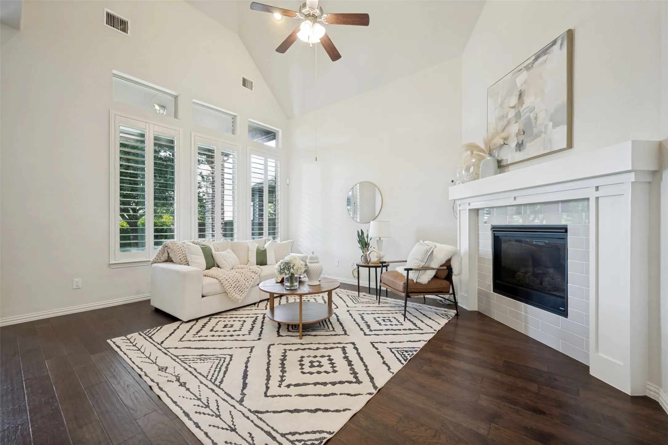 Vaulted ceiling with a ceiling fan, dark wood-finish flooring, white fireplace surround with a tile accent, large windows with white louvered shutters, and transom windows