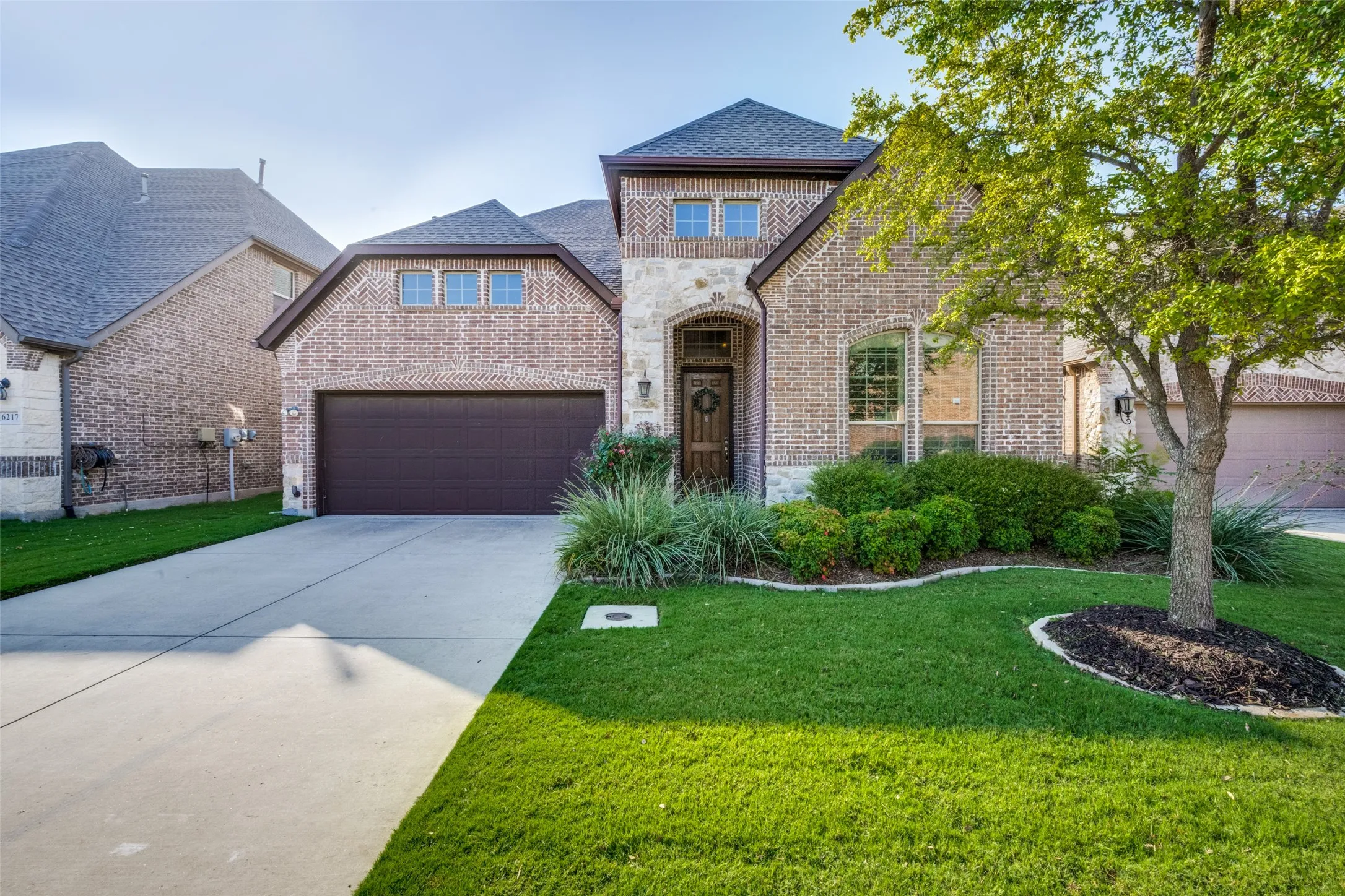 Brick and stone facade with an arched entryway, mature tree, concrete driveway, and established landscaping
