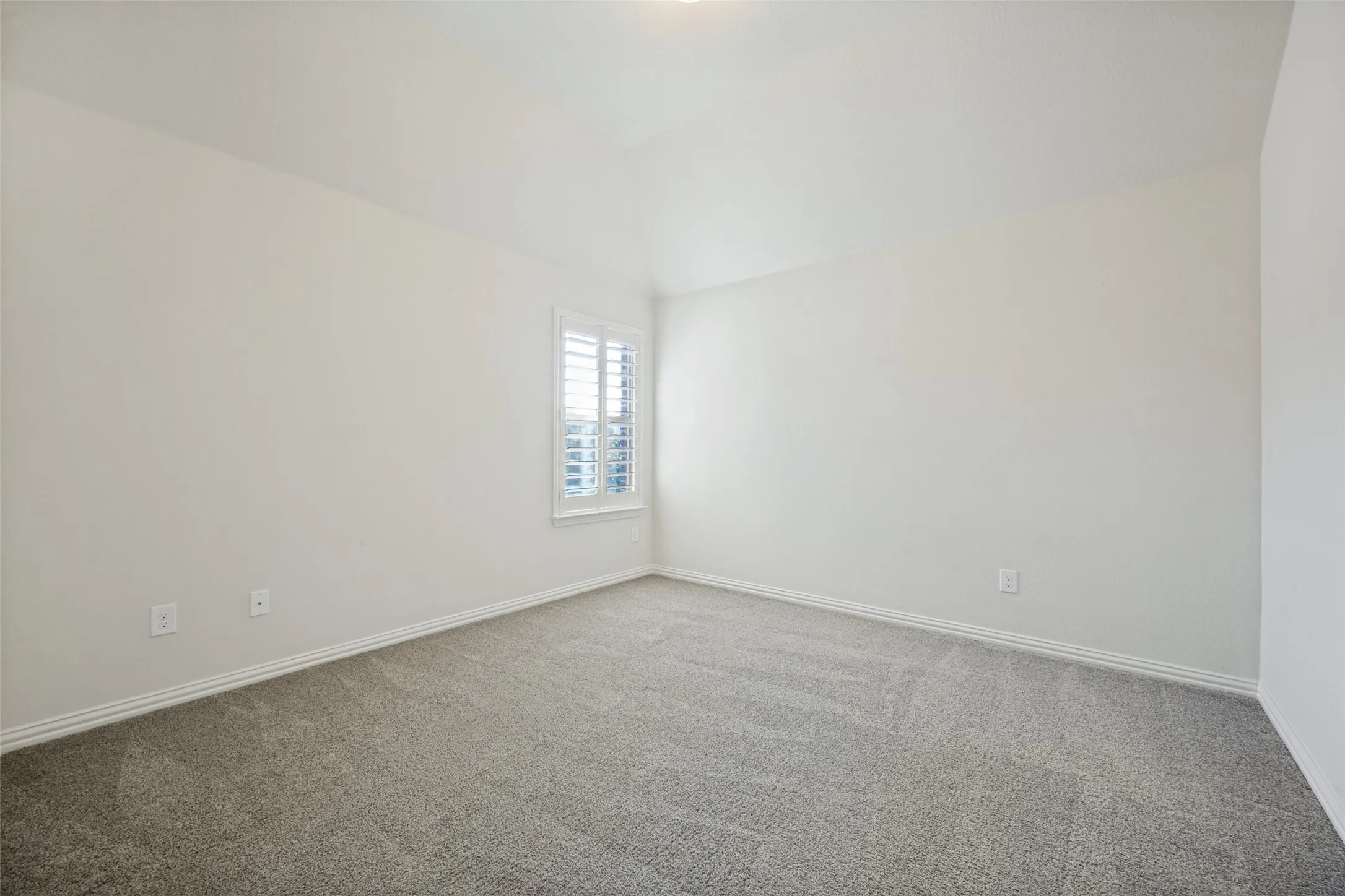 Light gray carpet flooring, white walls, vaulted ceiling, and a window with white louvered shutters