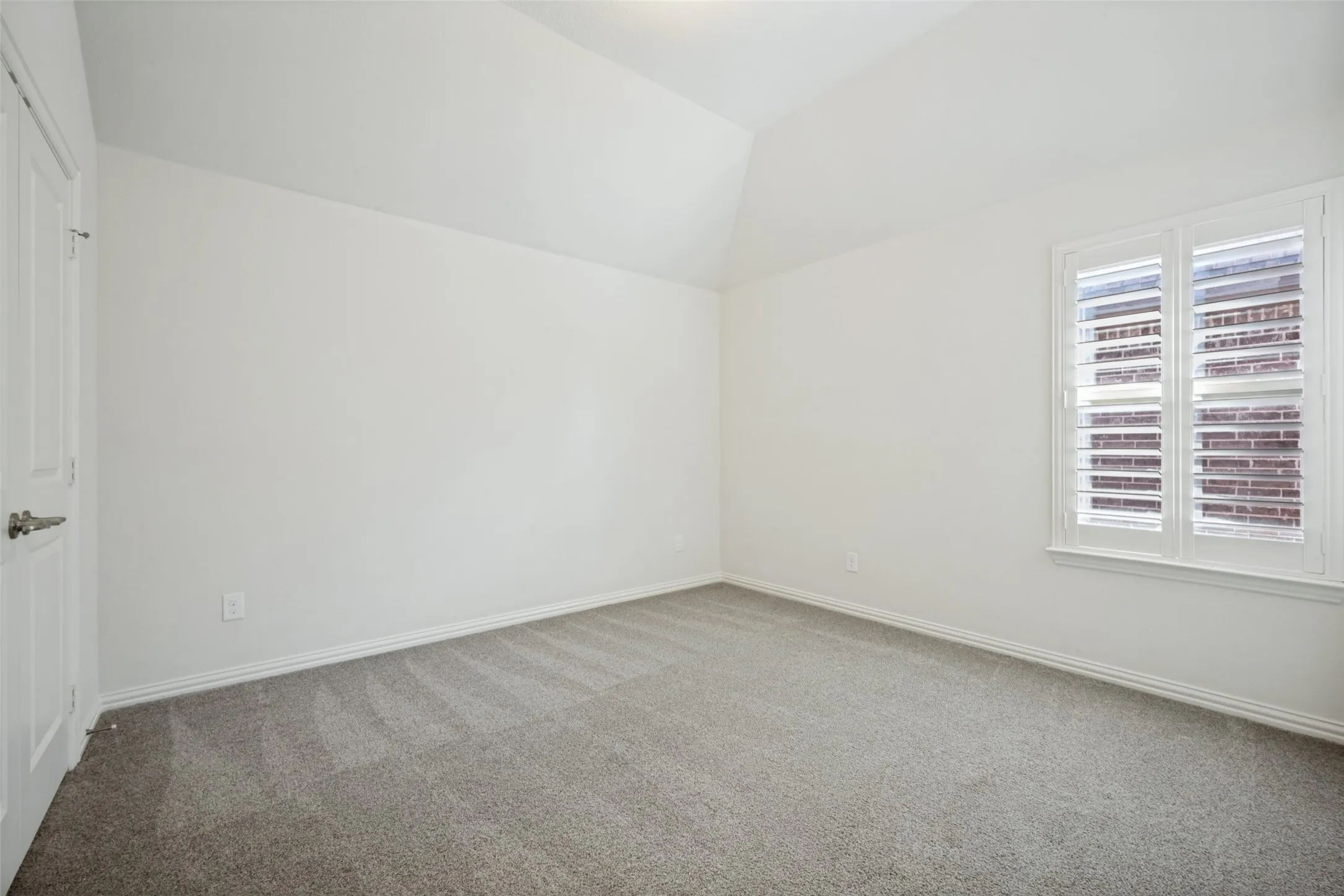 Vaulted ceiling, neutral wall paint, gray carpeting, white plantation shutters, and a white interior door with brushed nickel hardware