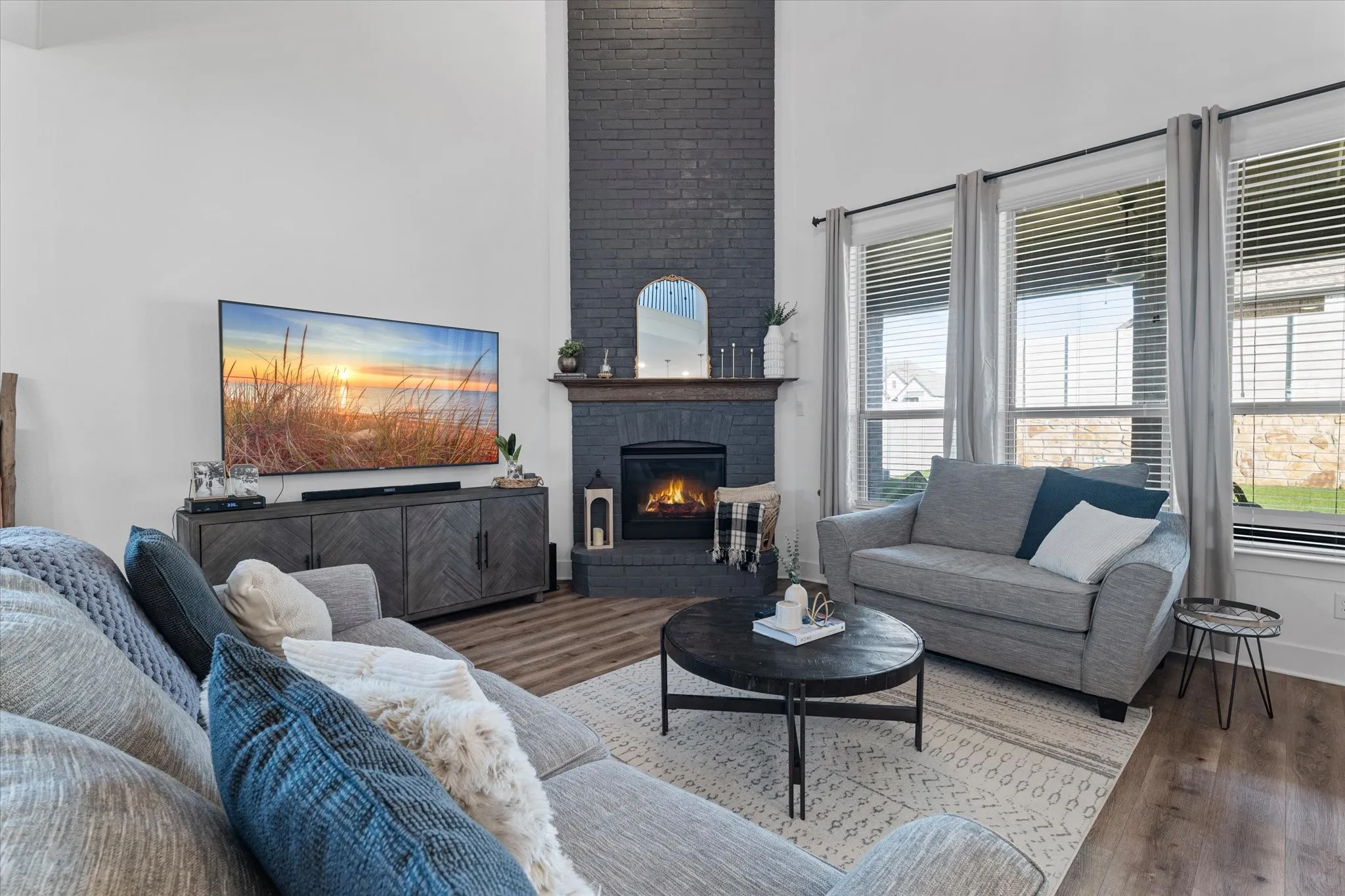 Vaulted ceiling, brick fireplace with wood mantel, built-in electric fireplace, wood-finish flooring, and multiple large windows