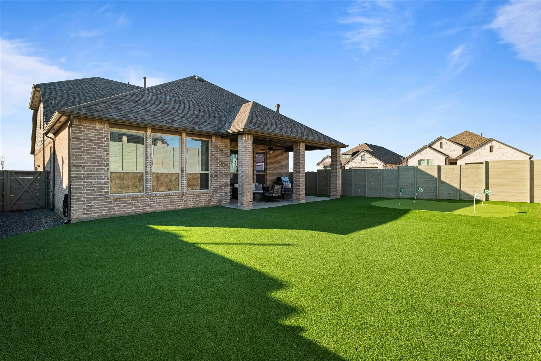 Expansive artificial turf backyard featuring a putting green, covered patio, brick exterior, and privacy fencing