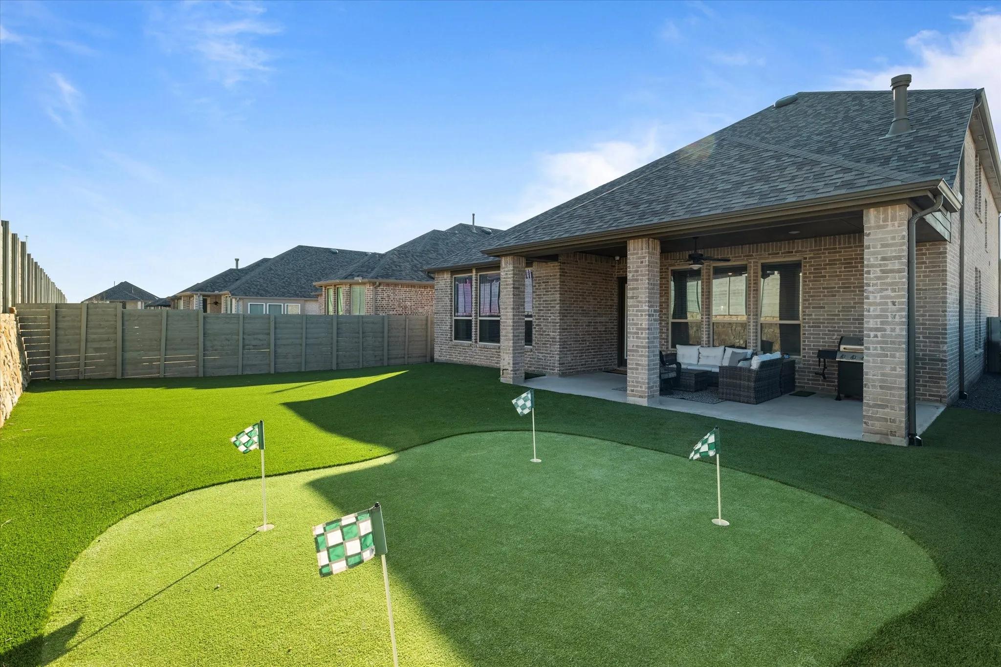 Expansive artificial turf backyard featuring a putting green with multiple holes, a covered patio with an outdoor ceiling fan, and a brick exterior