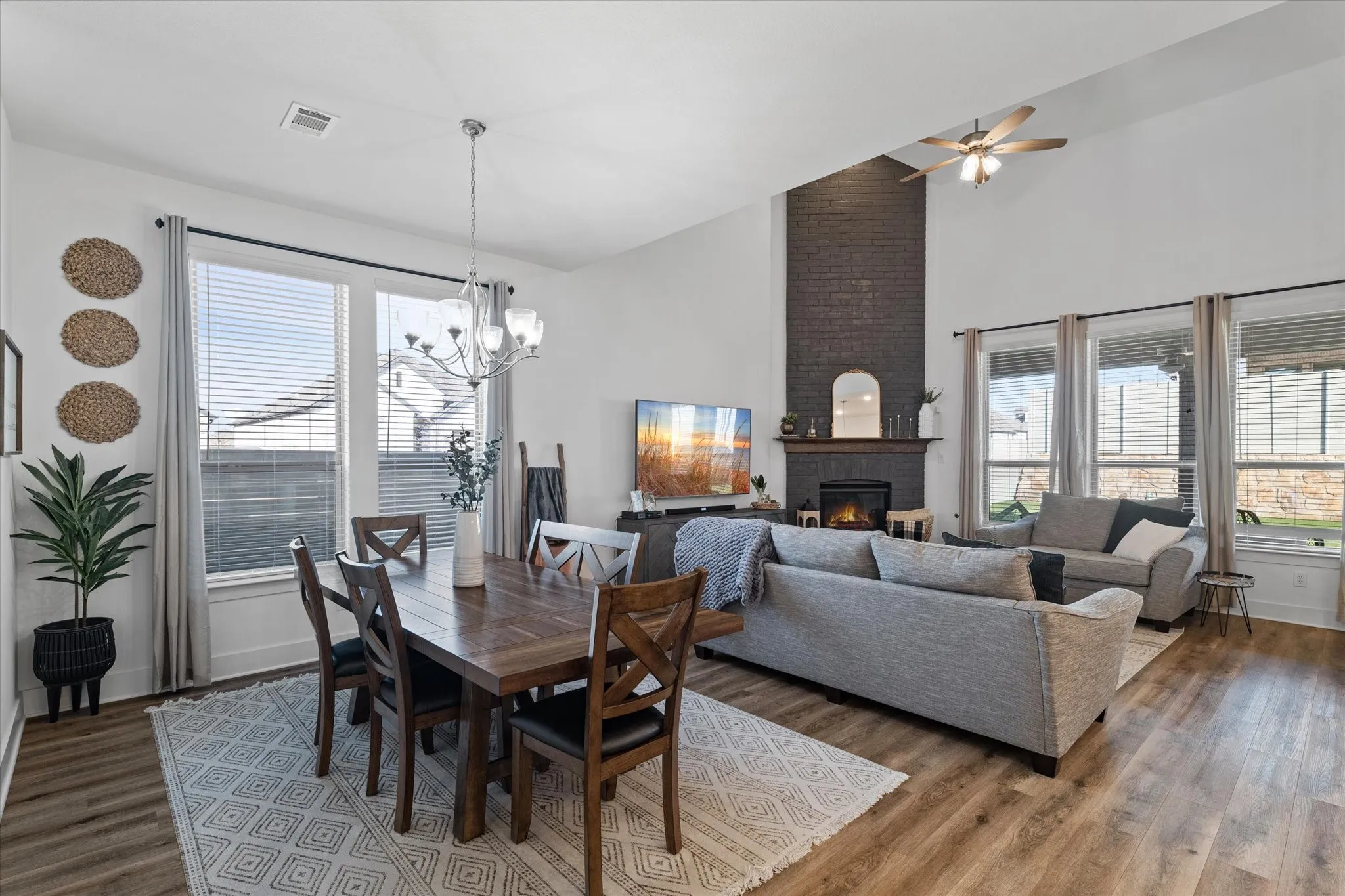 Vaulted ceiling with a ceiling fan, fireplace with a dark brick facade, wood-finish flooring, multiple windows with blinds, and a chandelier