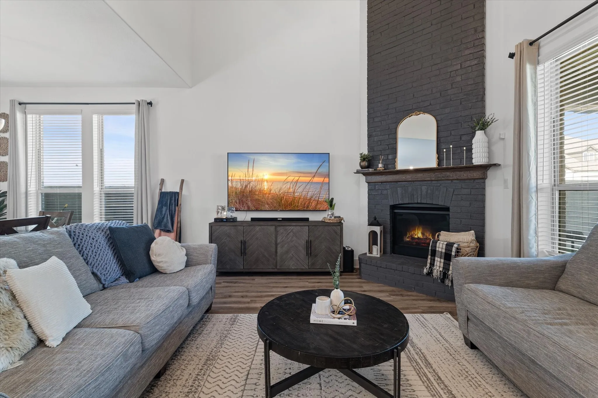 Vaulted ceiling, dark brick fireplace with a wood-finish mantel, wood-finish flooring, and large windows
