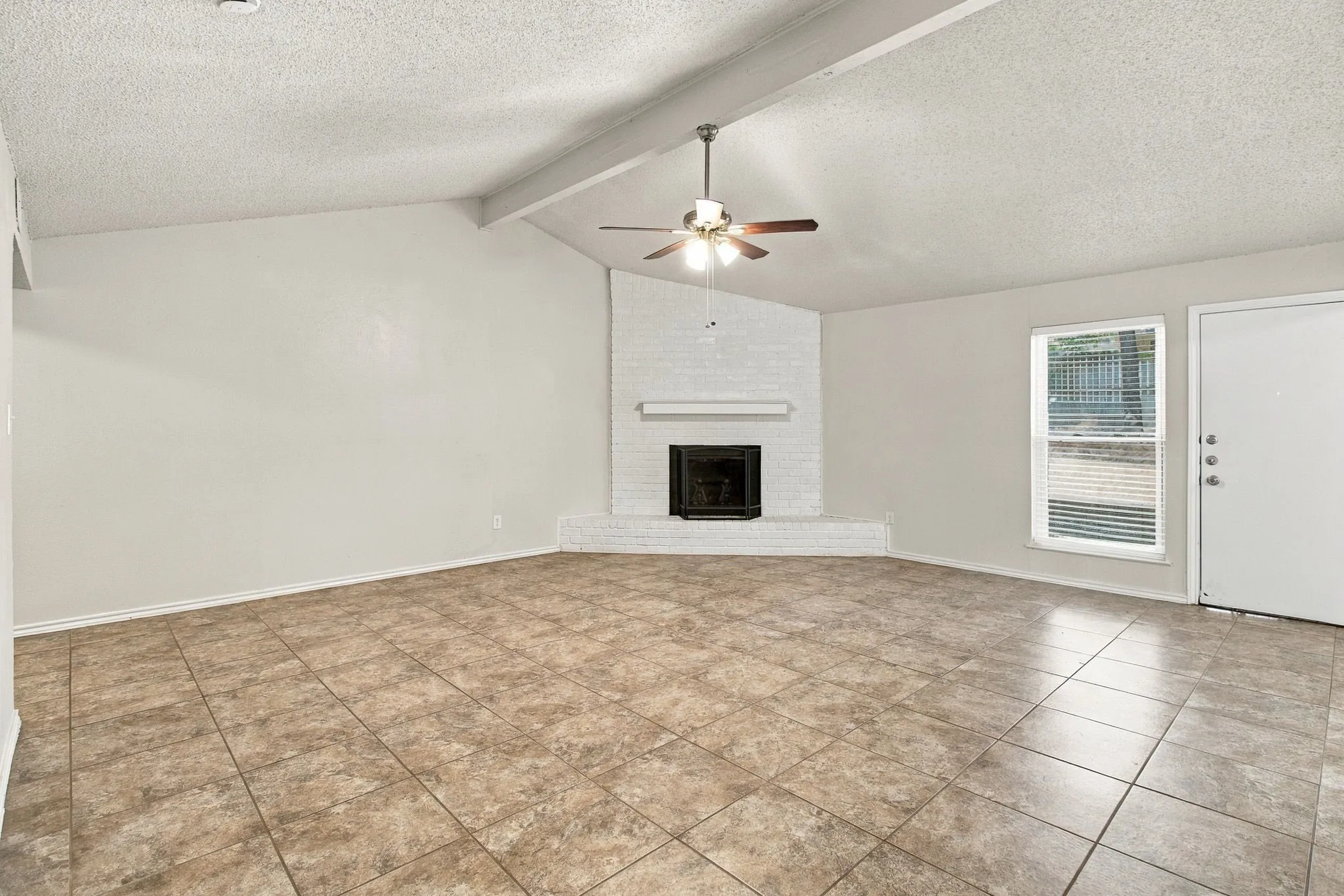 Vaulted ceiling with exposed beams, ceiling fan, white brick fireplace, tile flooring, and window with blinds