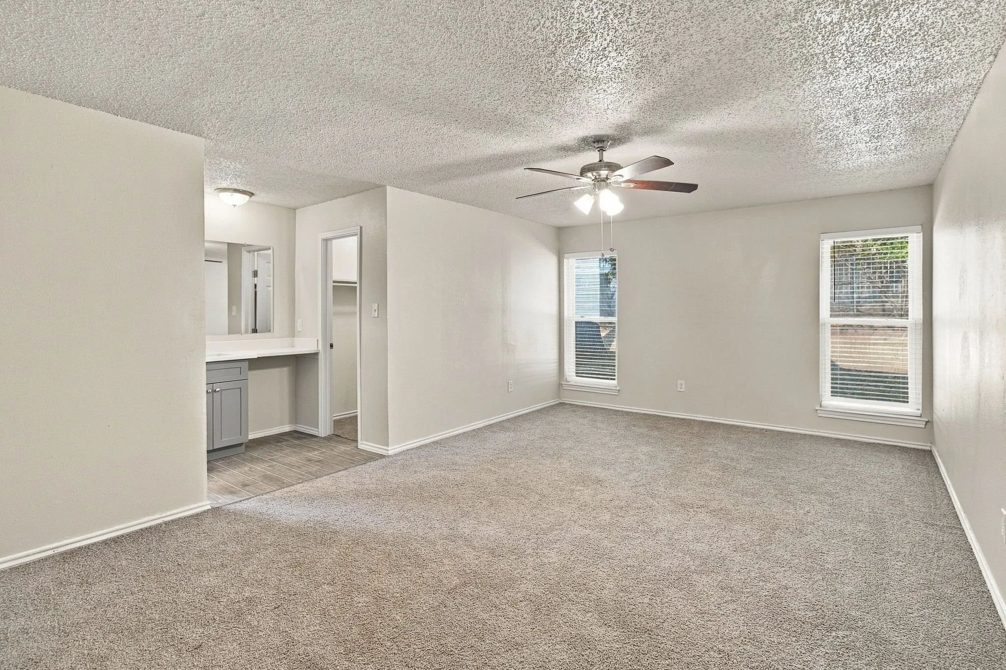 Spacious room with light-toned carpet flooring, two windows with blinds, and a ceiling fan
