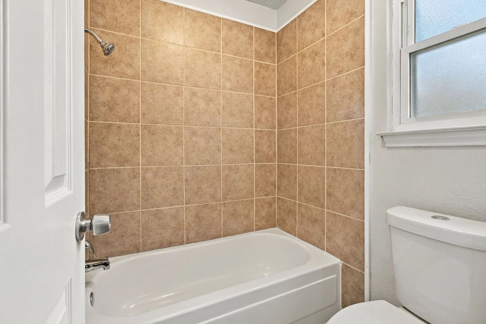 Bathtub and shower combination with tan tile surround, white toilet, and window with obscured glass