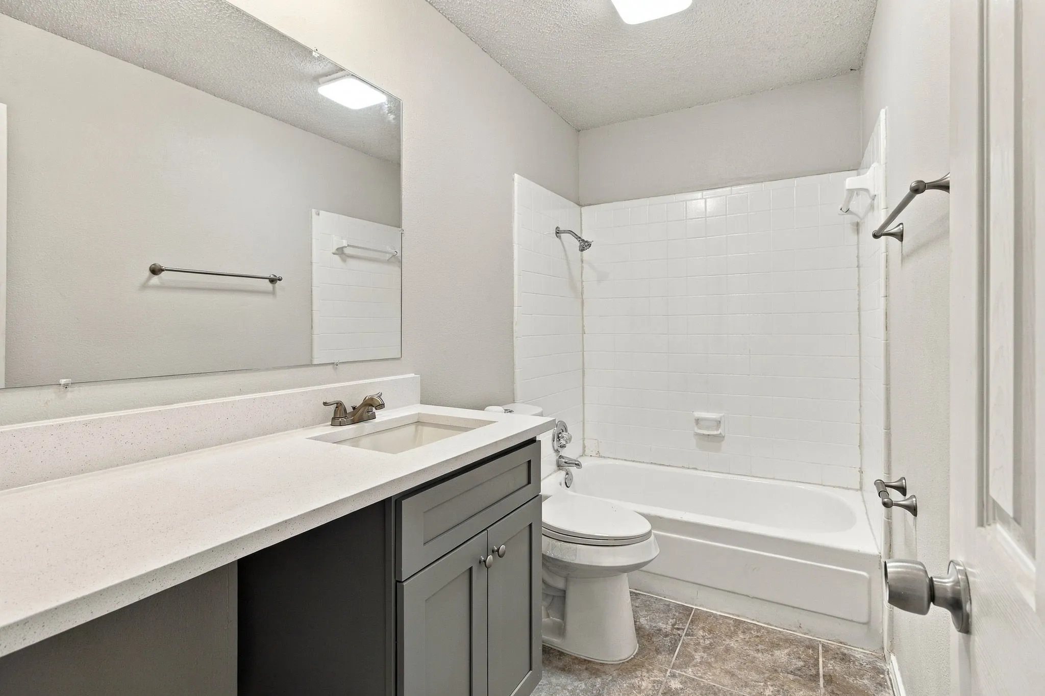 White countertop vanity with integrated sink, gray cabinetry, white tiled bathtub surround, and neutral floor tile