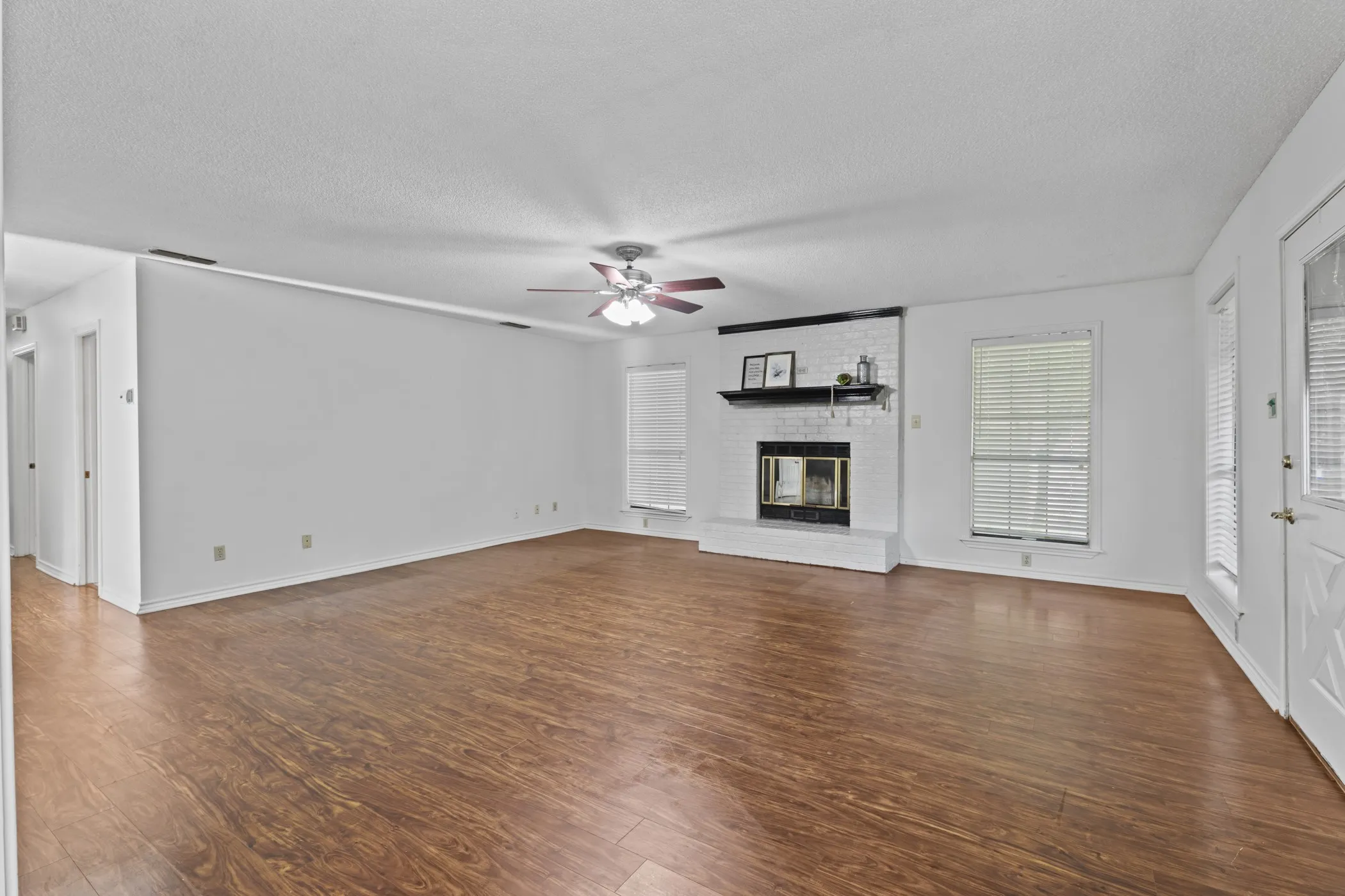 Expansive living area featuring wood-finish flooring, a white brick fireplace with a dark mantel, and multiple windows