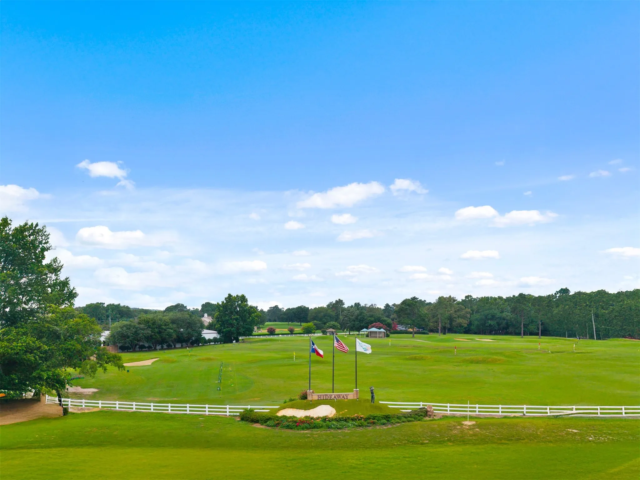 Expansive golf course setting featuring manicured greens, a decorative monument with flags, and a white fence border