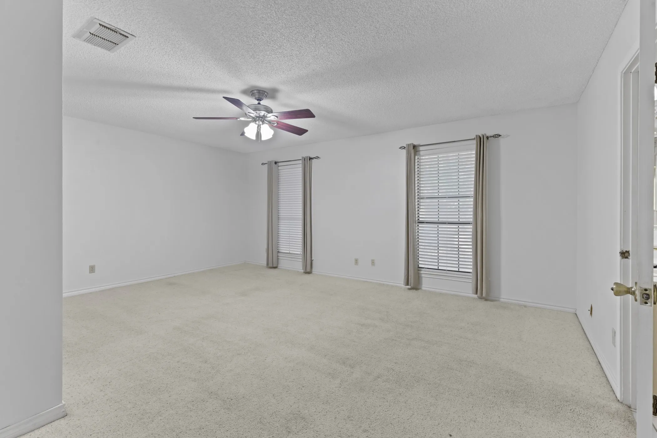 Expansive room featuring light-colored carpeting, two windows with blinds, a ceiling fan with integrated lighting, and a white paneled door