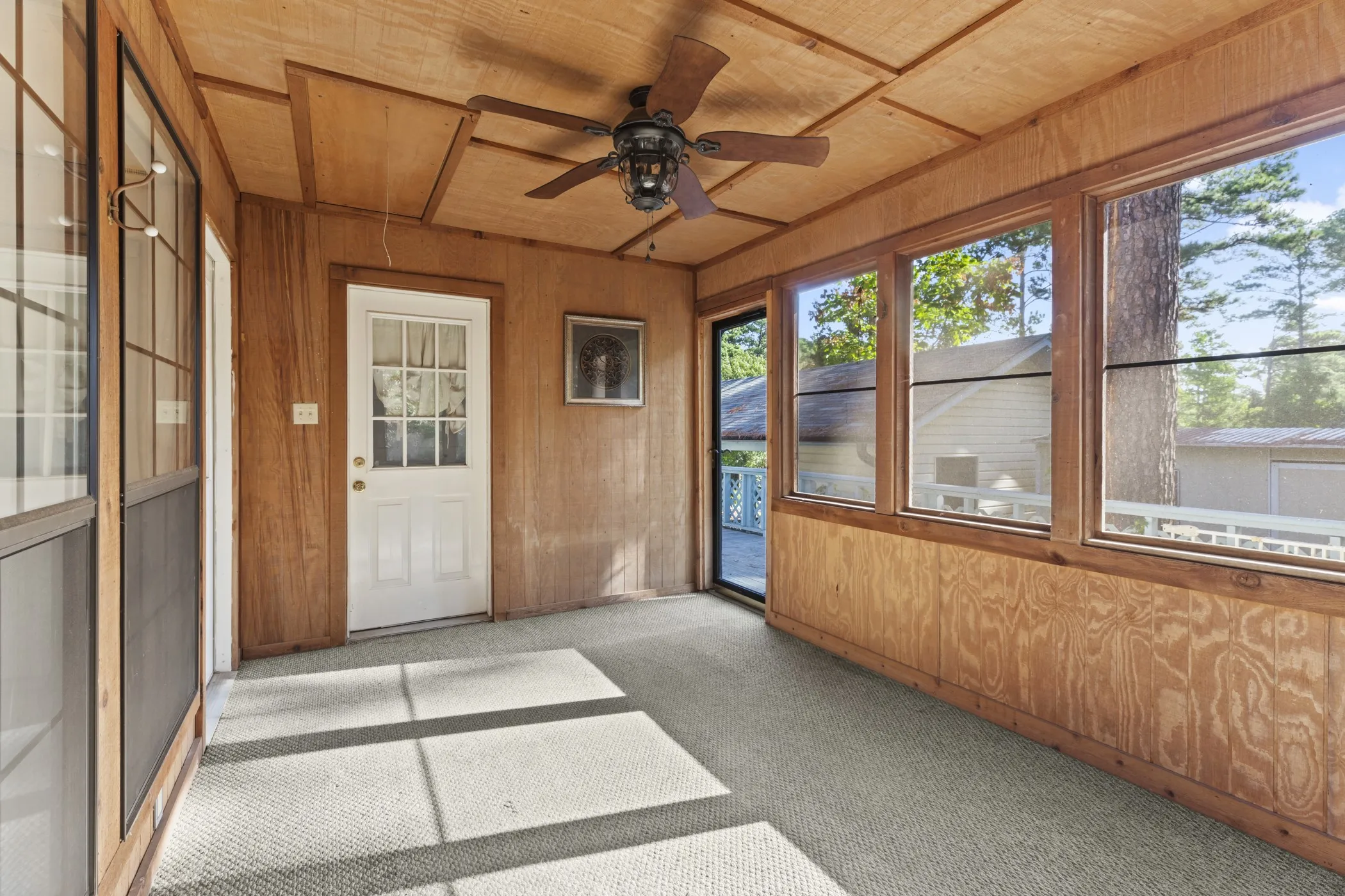 Enclosed porch featuring wood paneling, multiple large windows, a ceiling fan, a white exterior door, and light-colored carpeting