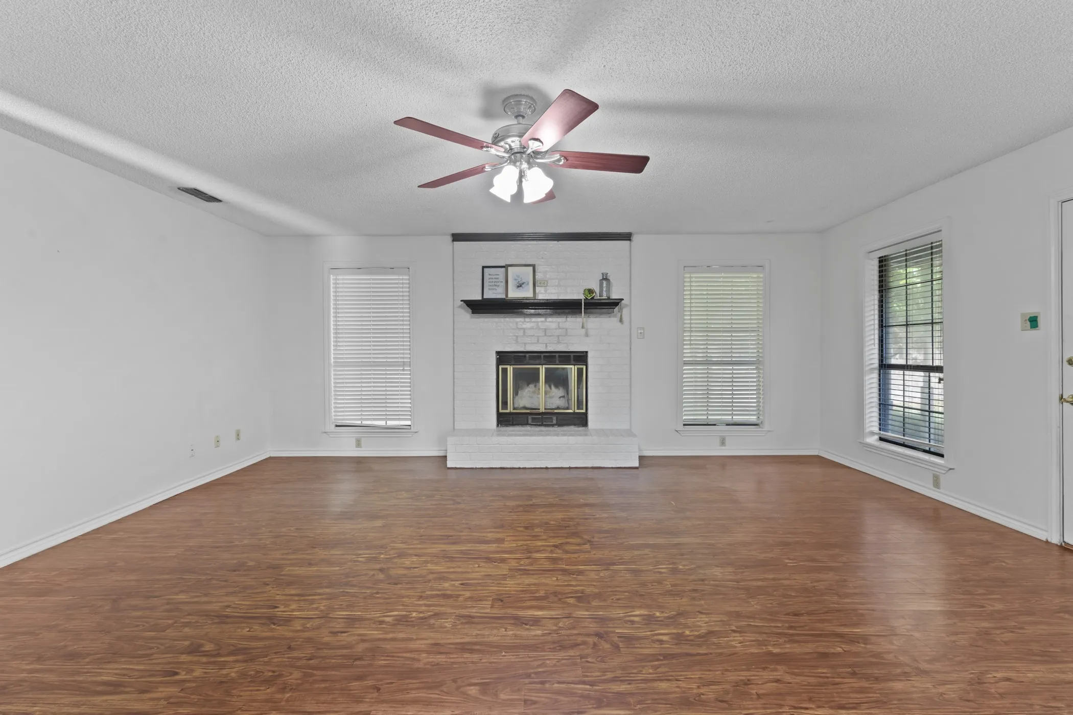 Spacious room featuring wood-finish flooring, a white brick fireplace, and multiple windows with blinds
