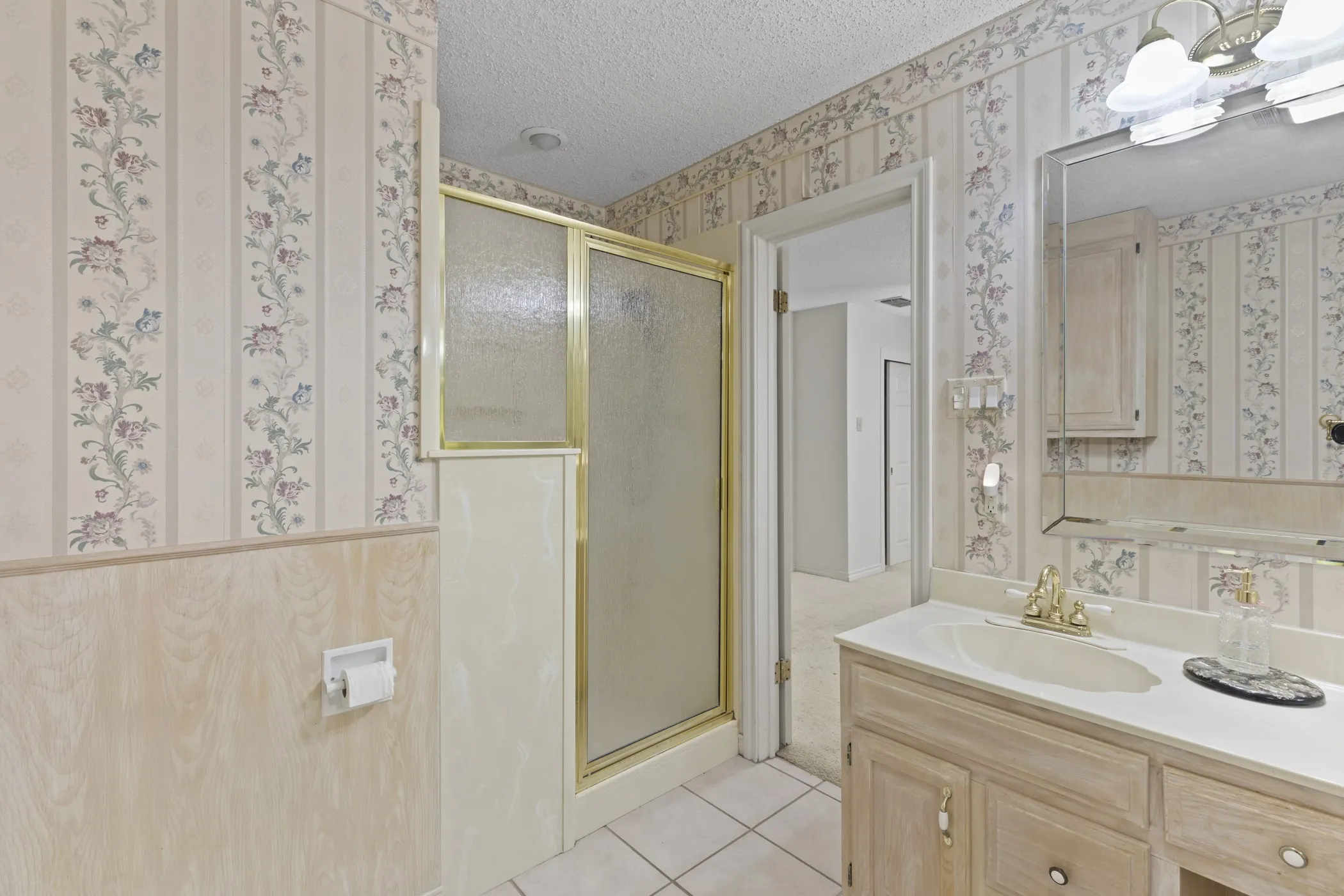 Bathroom featuring a vanity with an integrated sink, a mirrored medicine cabinet, a shower enclosure with a gold-tone frame, and floral wallpaper