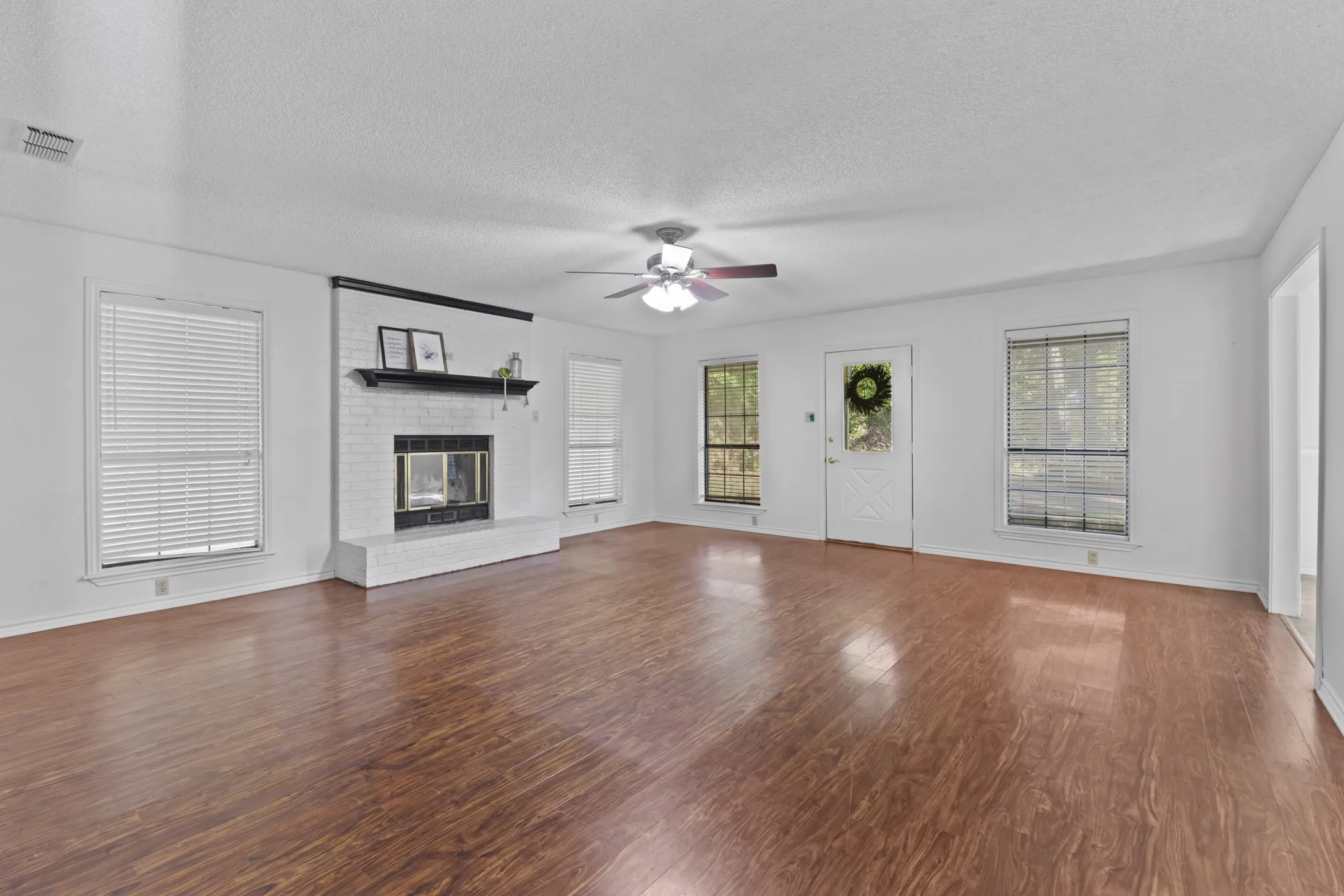 Spacious room featuring wood-finish flooring, a brick-surround fireplace with a mantel, multiple windows, and a ceiling fan with integrated lighting