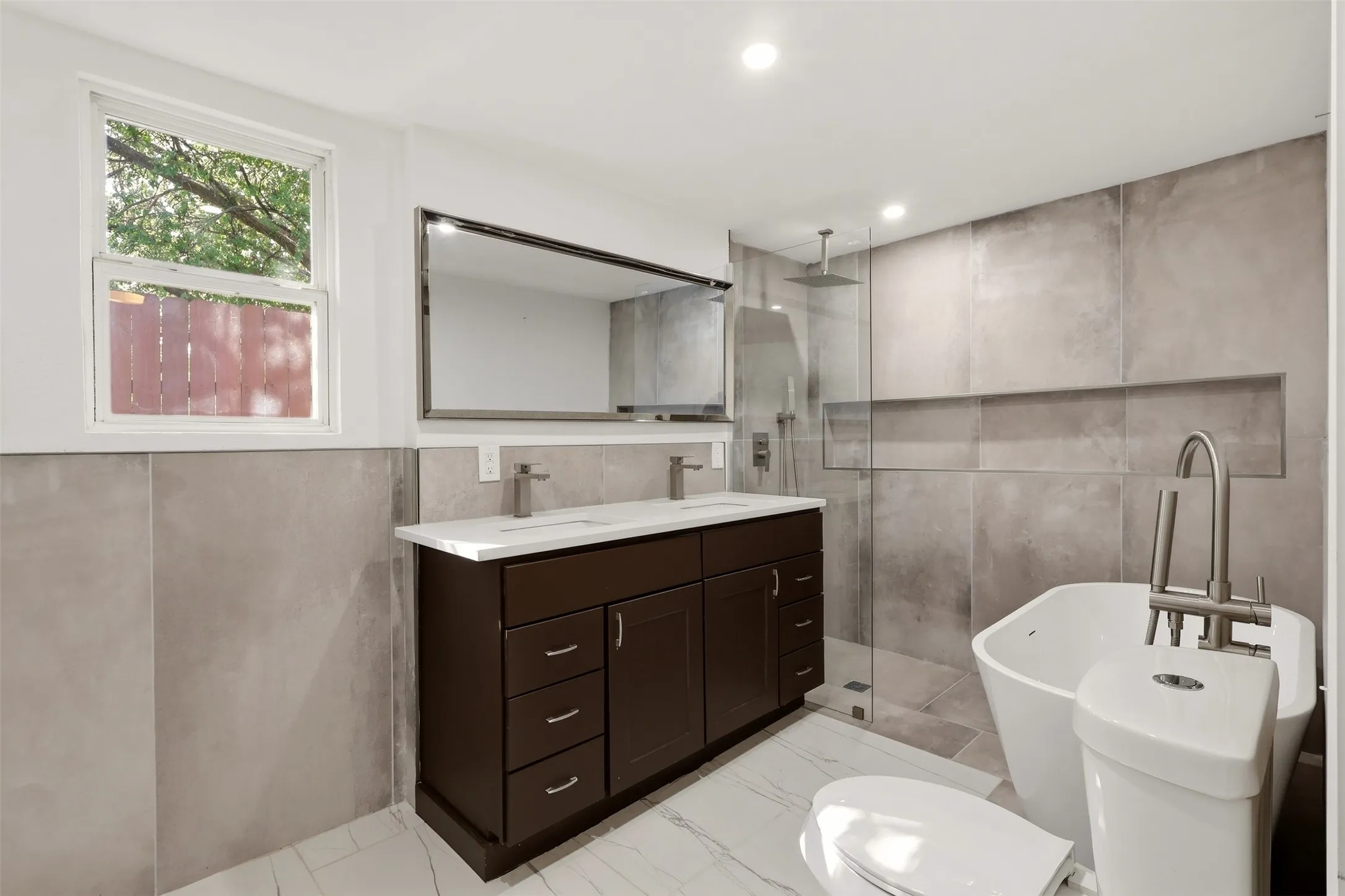 Double vanity with dual rectangular sinks and chrome fixtures, frameless glass shower enclosure with rainfall showerhead, freestanding soaking tub, recessed wall niche, and large format wall tiling