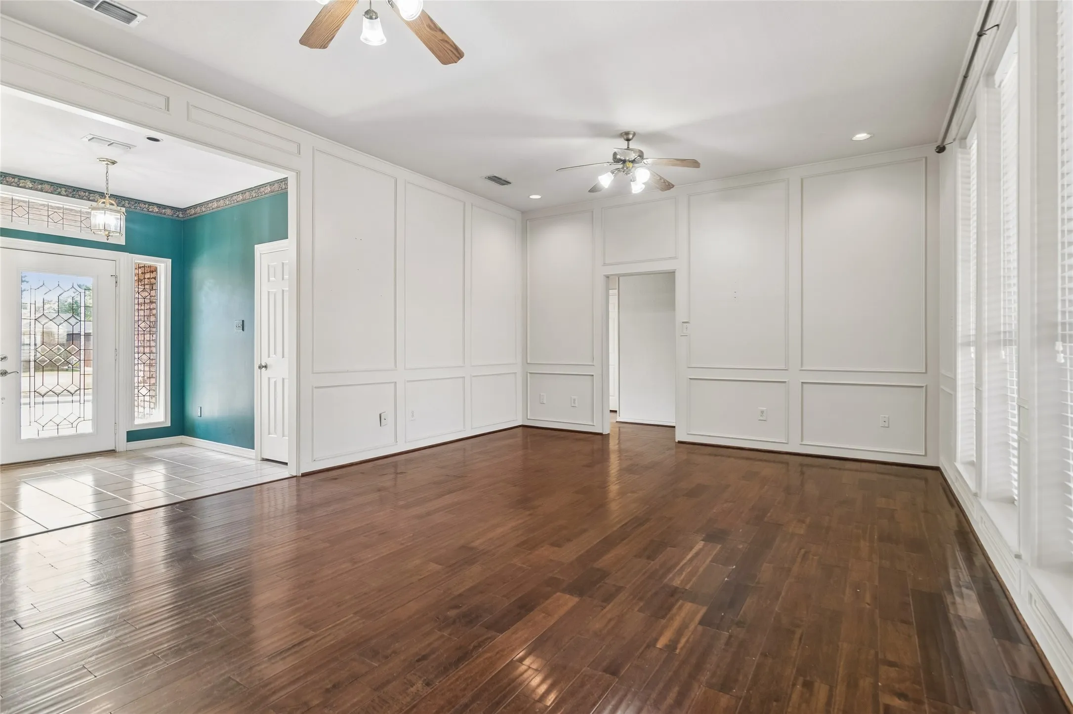 Wood-finish flooring throughout, white paneled walls, multiple ceiling fans, recessed lighting, and large windows with blinds
