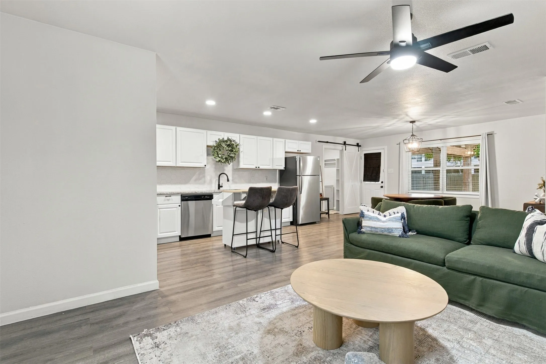 Open-concept living area featuring wood-finish flooring, white cabinetry, stainless steel appliances, and a modern ceiling fan