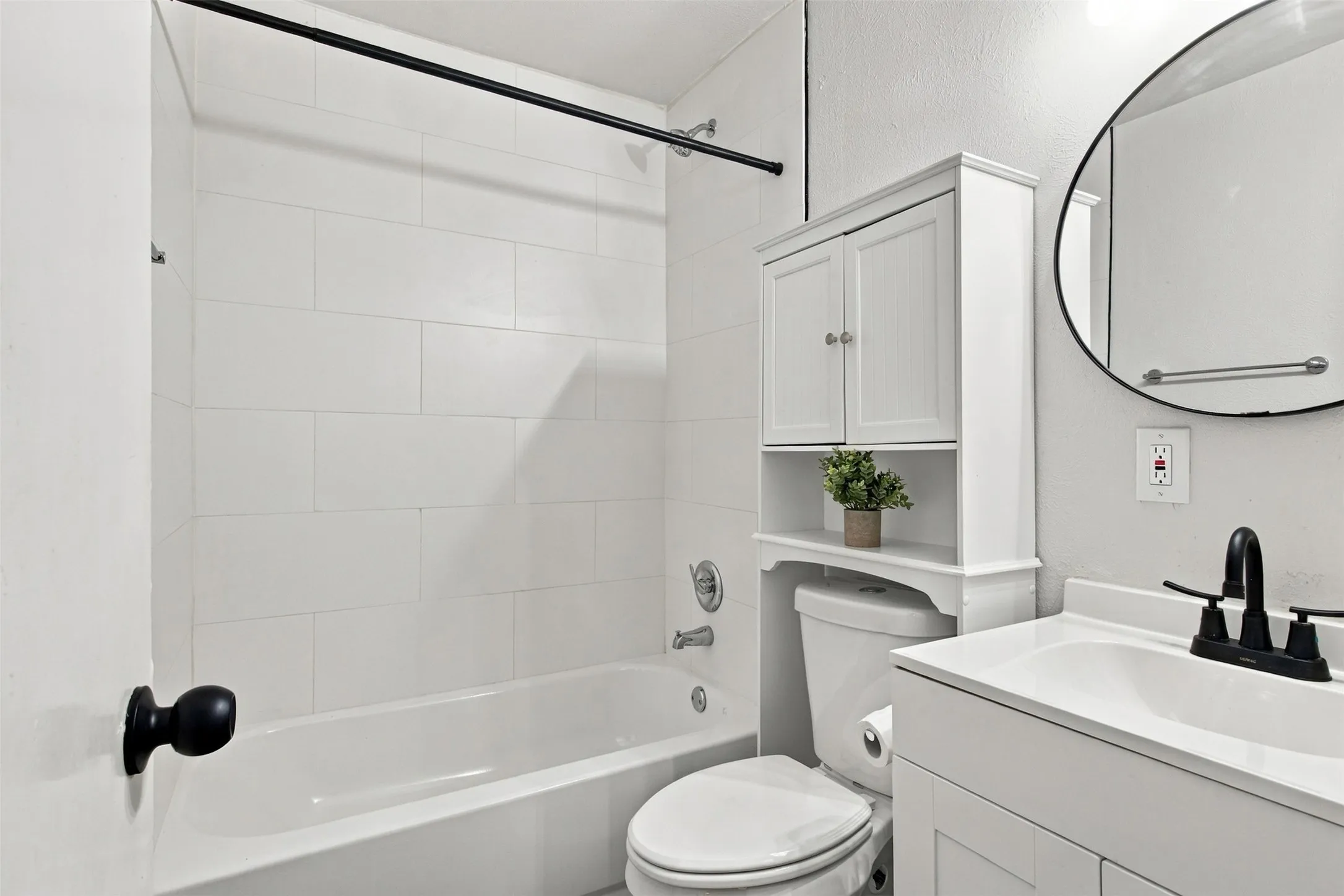 White tiled bathtub with a black shower rod, white vanity with an integrated sink, black two-handle faucet, circular mirror, and an over-the-toilet storage cabinet