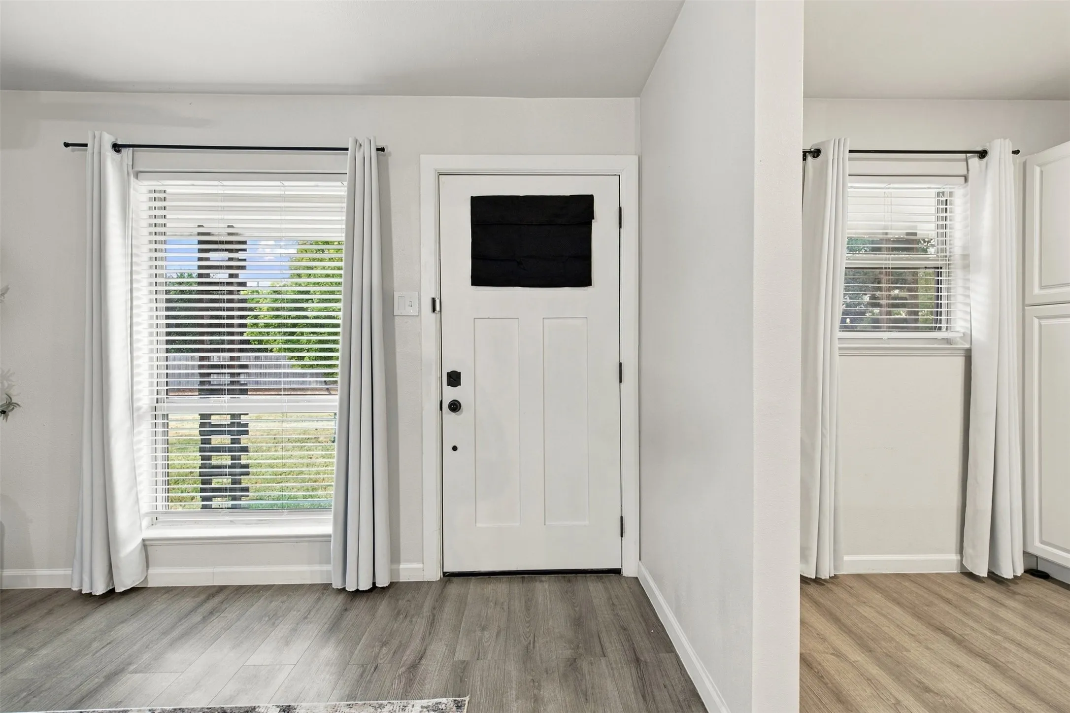 Wood-finish flooring throughout, white walls, white paneled door with dark hardware, and white window blinds