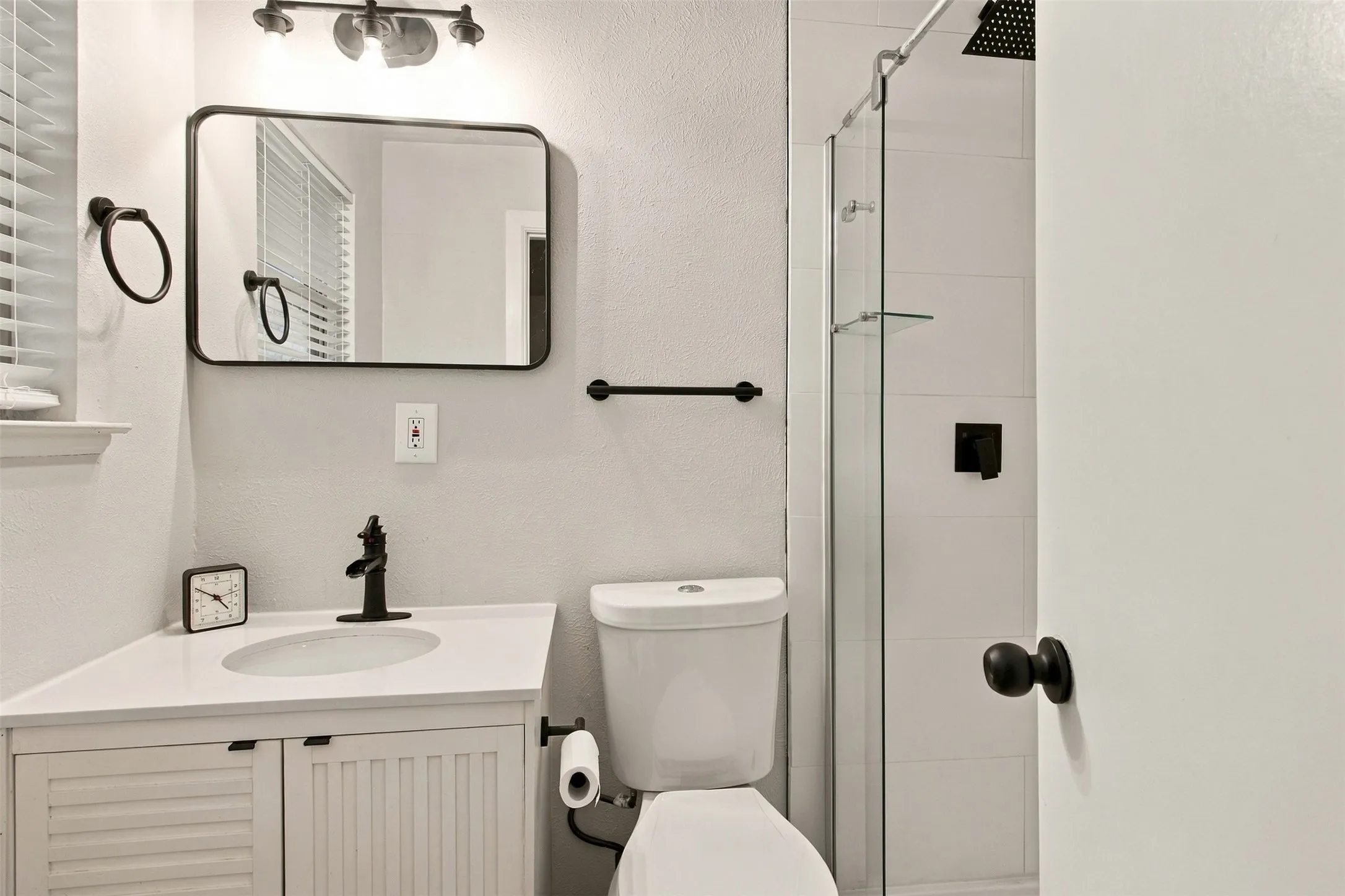 Corner vanity with a single basin and matte black faucet, glass-enclosed shower with black fixtures, white tile shower surround, and a rectangular framed mirror
