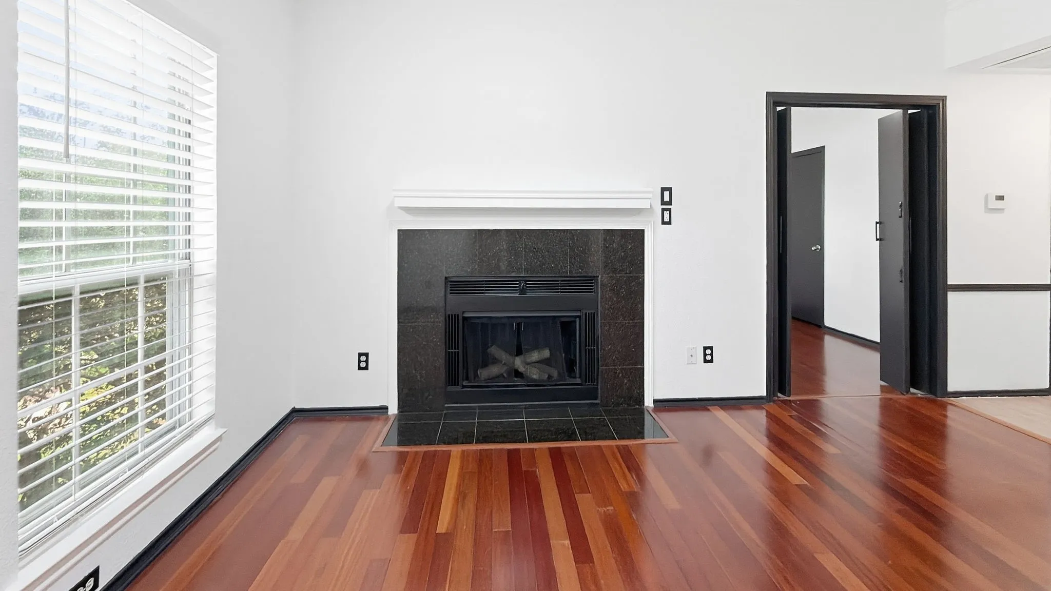 Hardwood flooring, black tile fireplace surround, white mantel, and a window with blinds